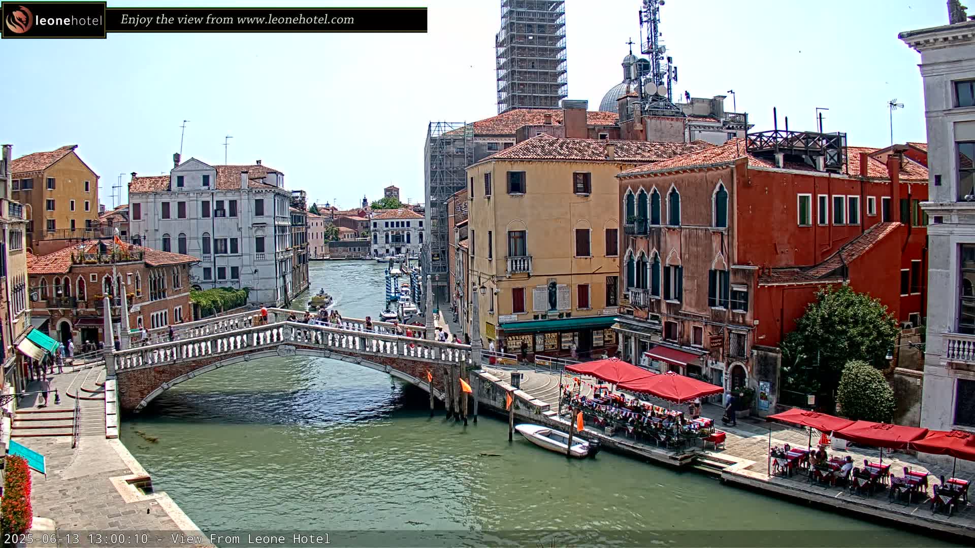 A sunny day in Venice shows a canal with a stone bridge, colorful buildings lining the waterway, and several outdoor cafes with red umbrellas.