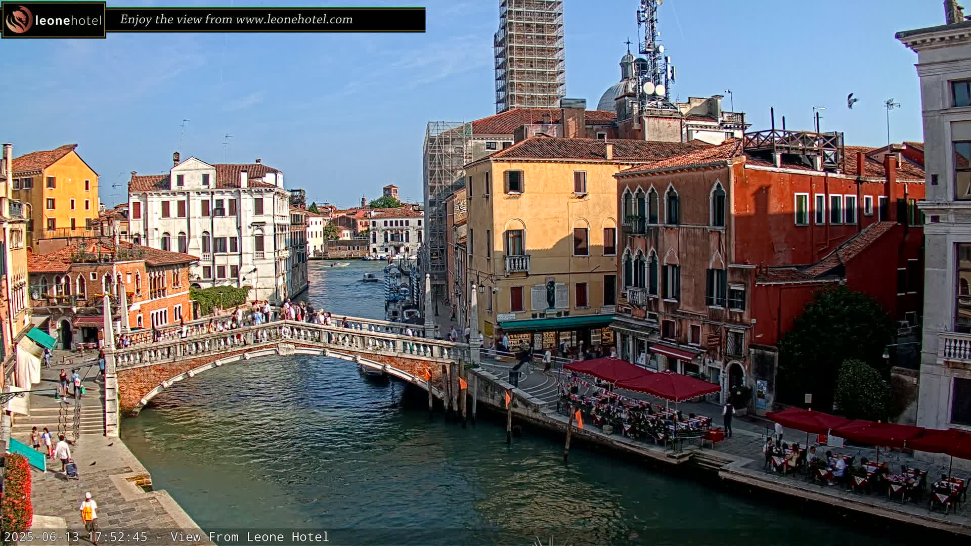 A sunny day in Venice shows a canal with a stone bridge, colorful buildings lining the waterway, and outdoor seating at restaurants.
