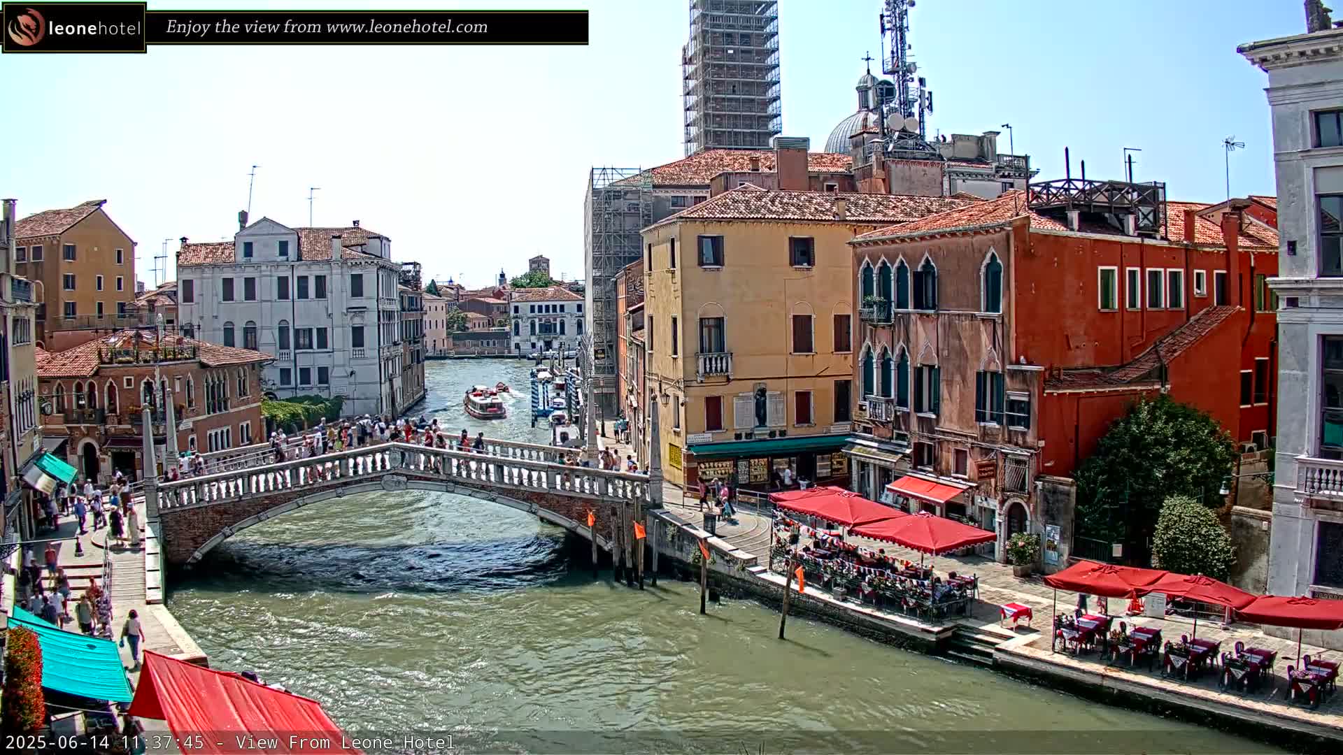 A sunny day in Venice shows a canal with a stone bridge crowded with people, boats on the water, and colorful buildings lining the waterway, some with outdoor cafes.