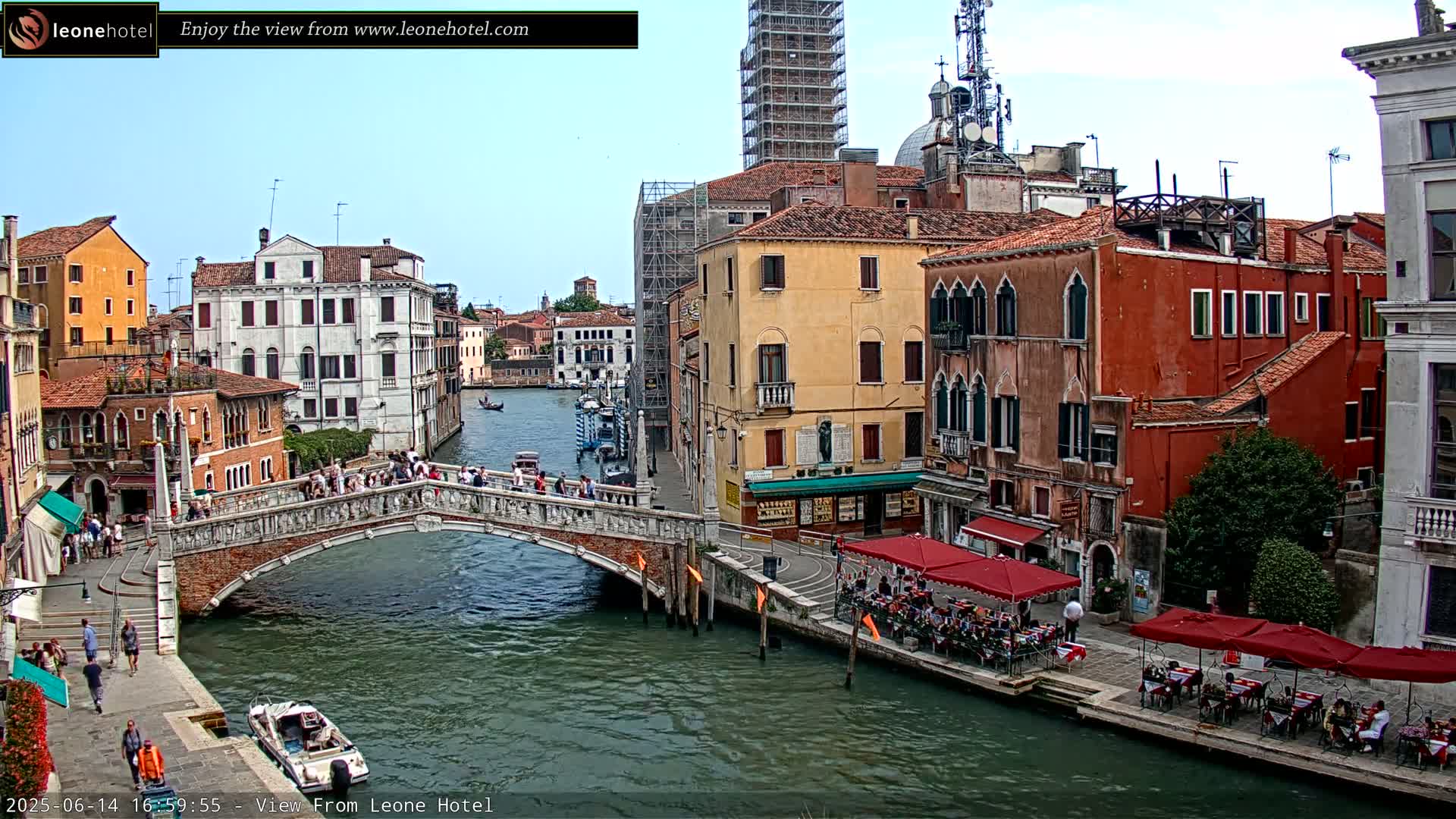 A canal in Venice, Italy, on a sunny day, shows a stone bridge with pedestrians crossing, boats on the water, and waterfront cafes with patrons seated outdoors.