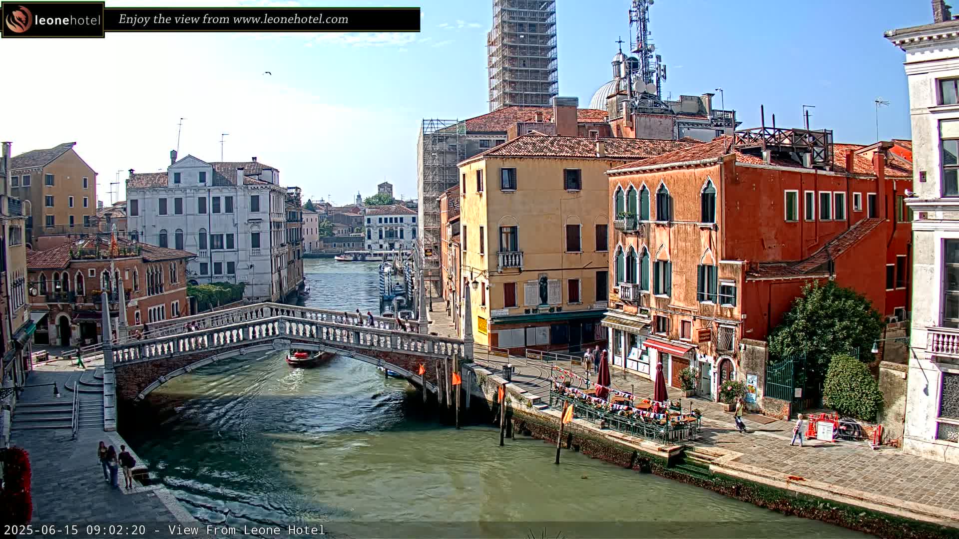 A sunny day in Venice shows a canal with a stone bridge, colorful buildings lining the waterway, and a few pedestrians walking along the canal's edge.
