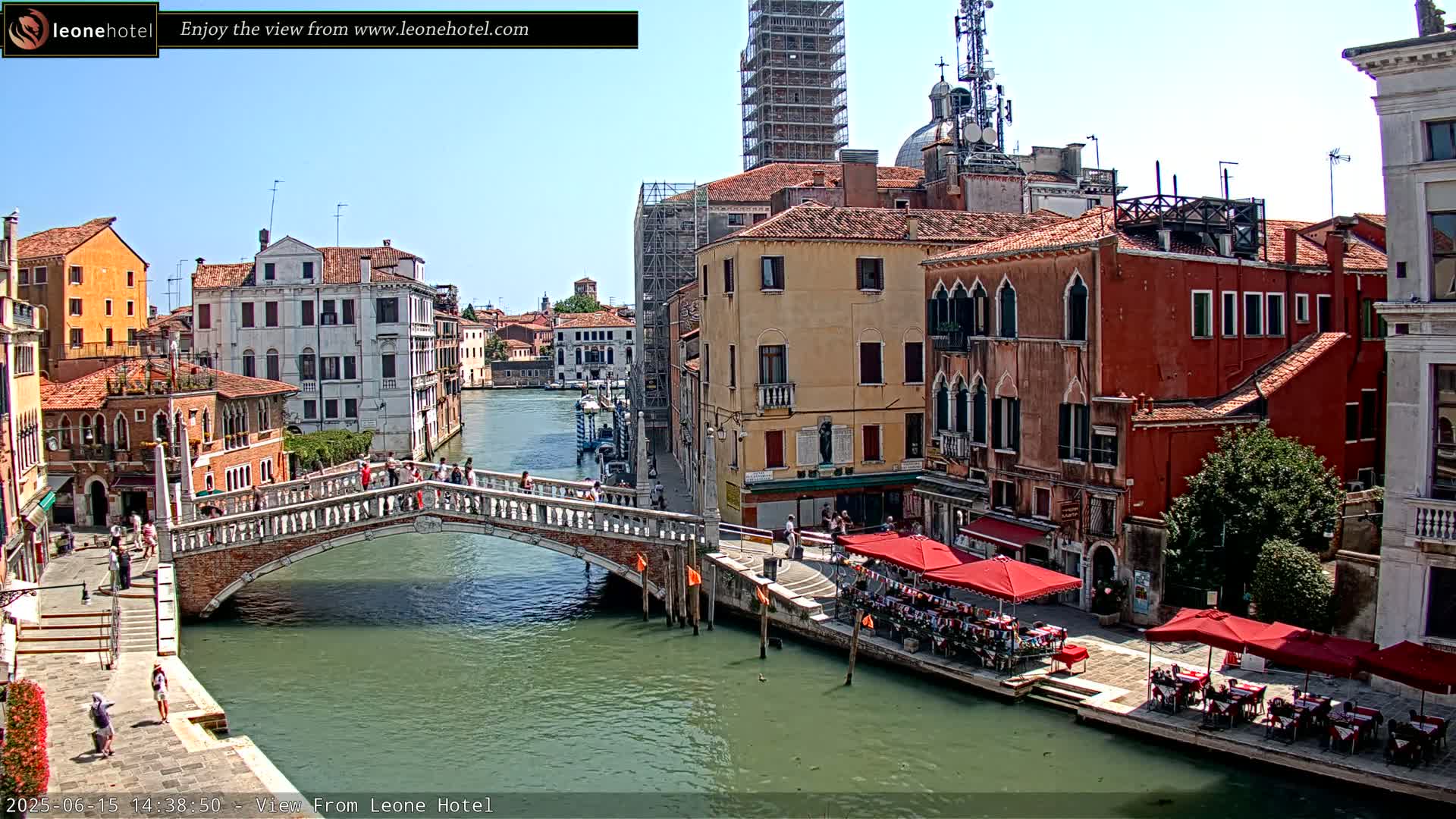 A sunny day in Venice shows a canal with a stone bridge, several colorful buildings lining its banks, and outdoor seating areas at restaurants.