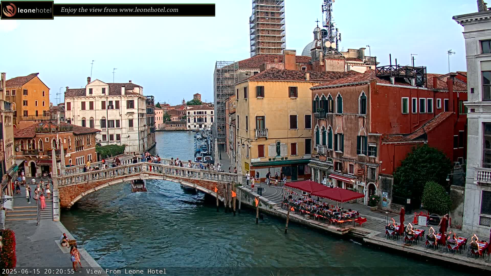 A bridge over a canal in Venice, Italy, with several people walking across it and others sitting at outdoor restaurants along the water's edge under a clear sky.