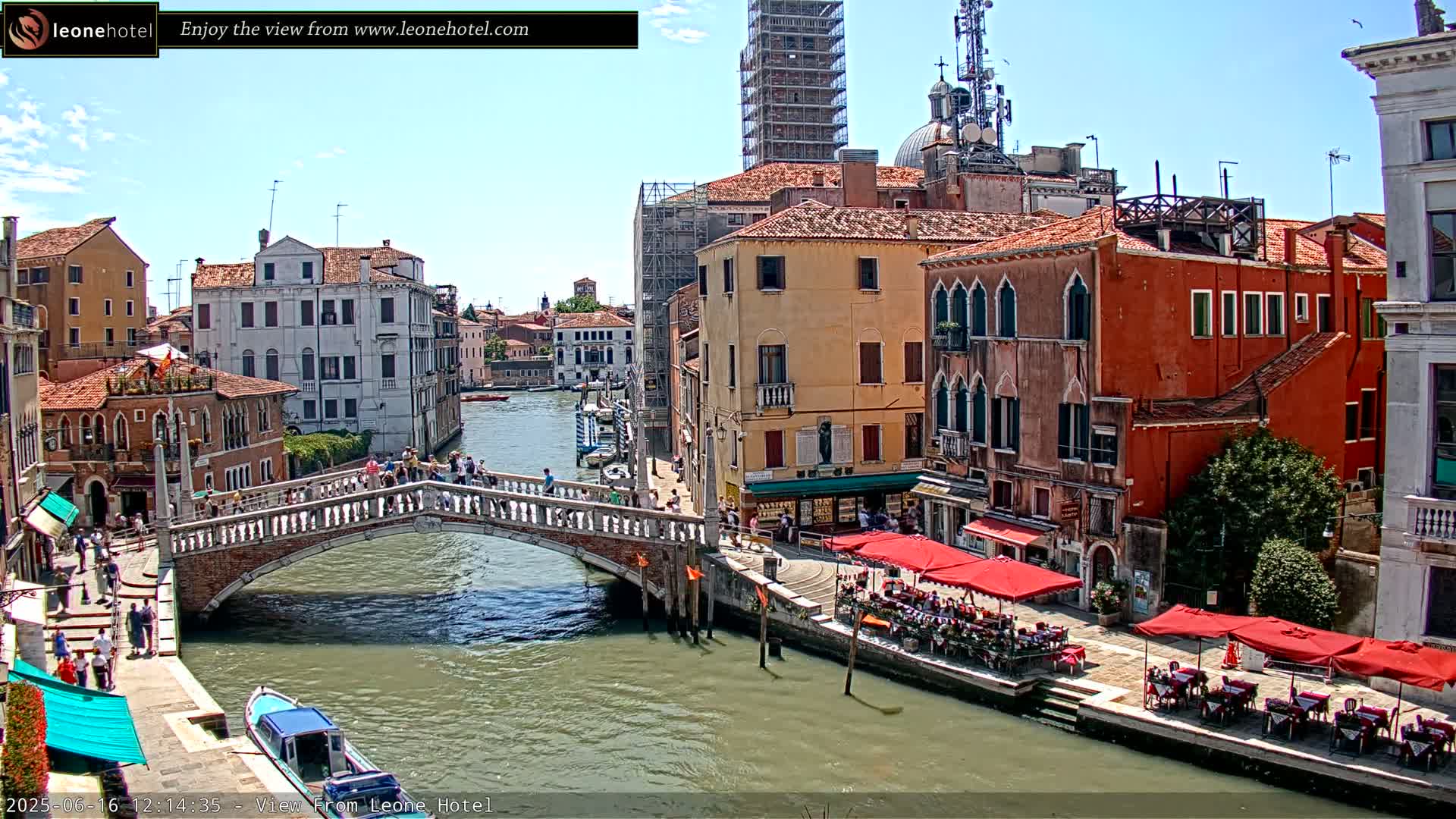 A sunny day in Venice shows a canal with a bridge, colorful buildings lining the waterway, and outdoor seating at cafes.