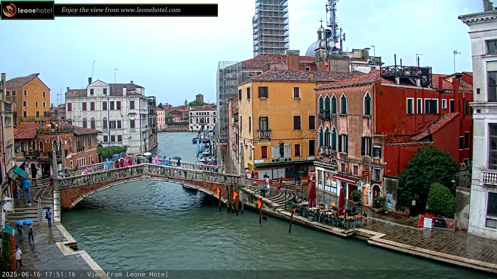 A rain-soaked canal in Venice, Italy, is viewed from a bridge, with colorful buildings lining the waterway and several people with umbrellas walking along the bridge and quay.