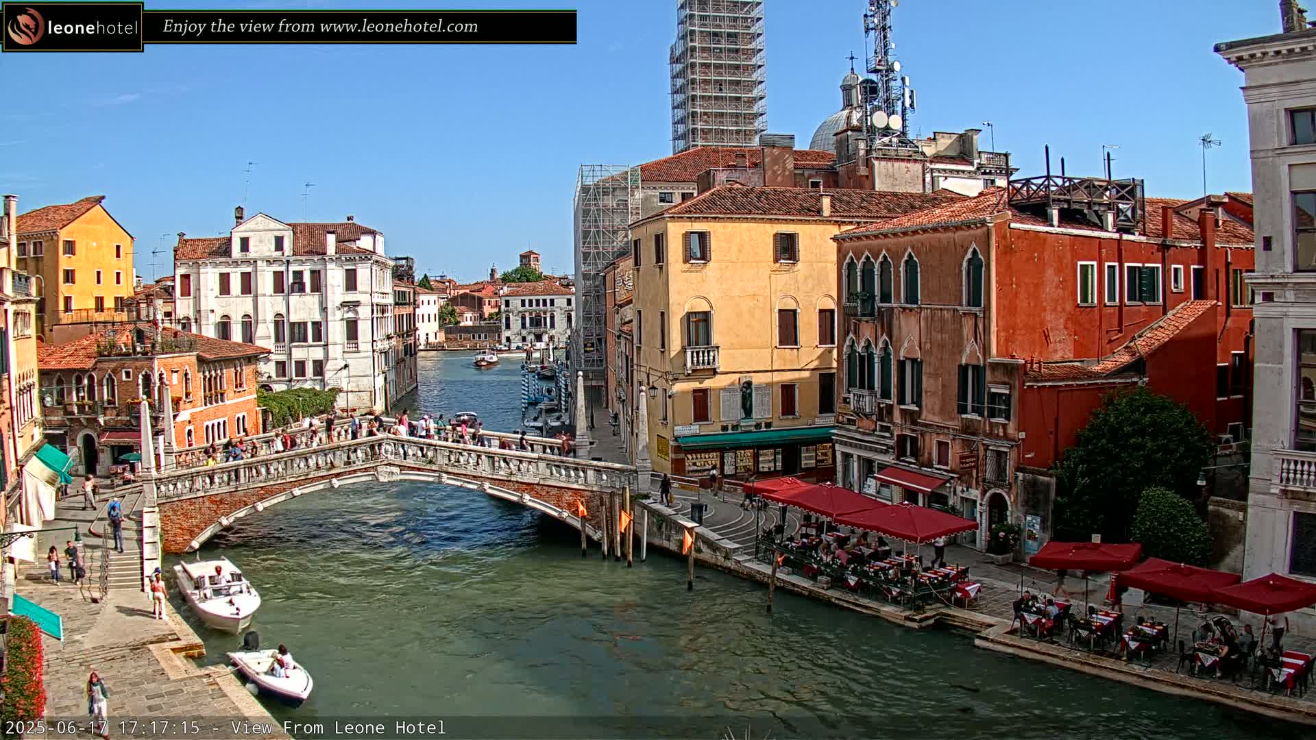 A sunny day in Venice shows a canal with a stone bridge crowded with people, boats on the water, and colorful buildings lining the waterway, with some buildings under construction.