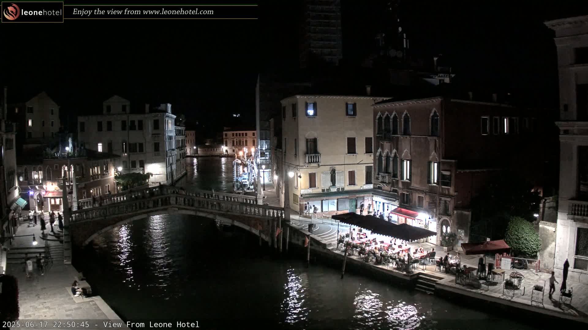 A nighttime view of a canal in Venice, Italy, showing a bridge, buildings, and an outdoor restaurant with people dining under a canopy.