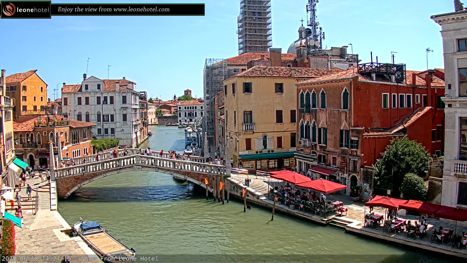 A sunny day in Venice shows a canal with a stone bridge, colorful buildings lining the waterway, and outdoor seating at cafes along the banks.