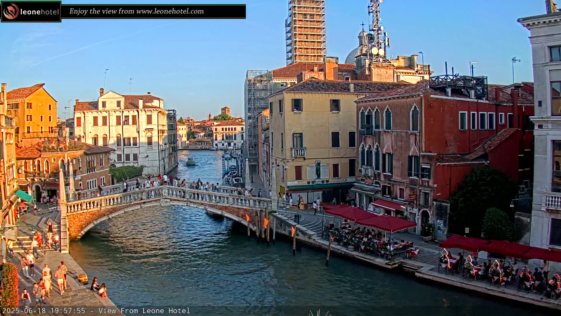 A bridge spans a canal in Venice, Italy, under a clear evening sky, with pedestrians walking across and people dining at outdoor cafes alongside the waterway.