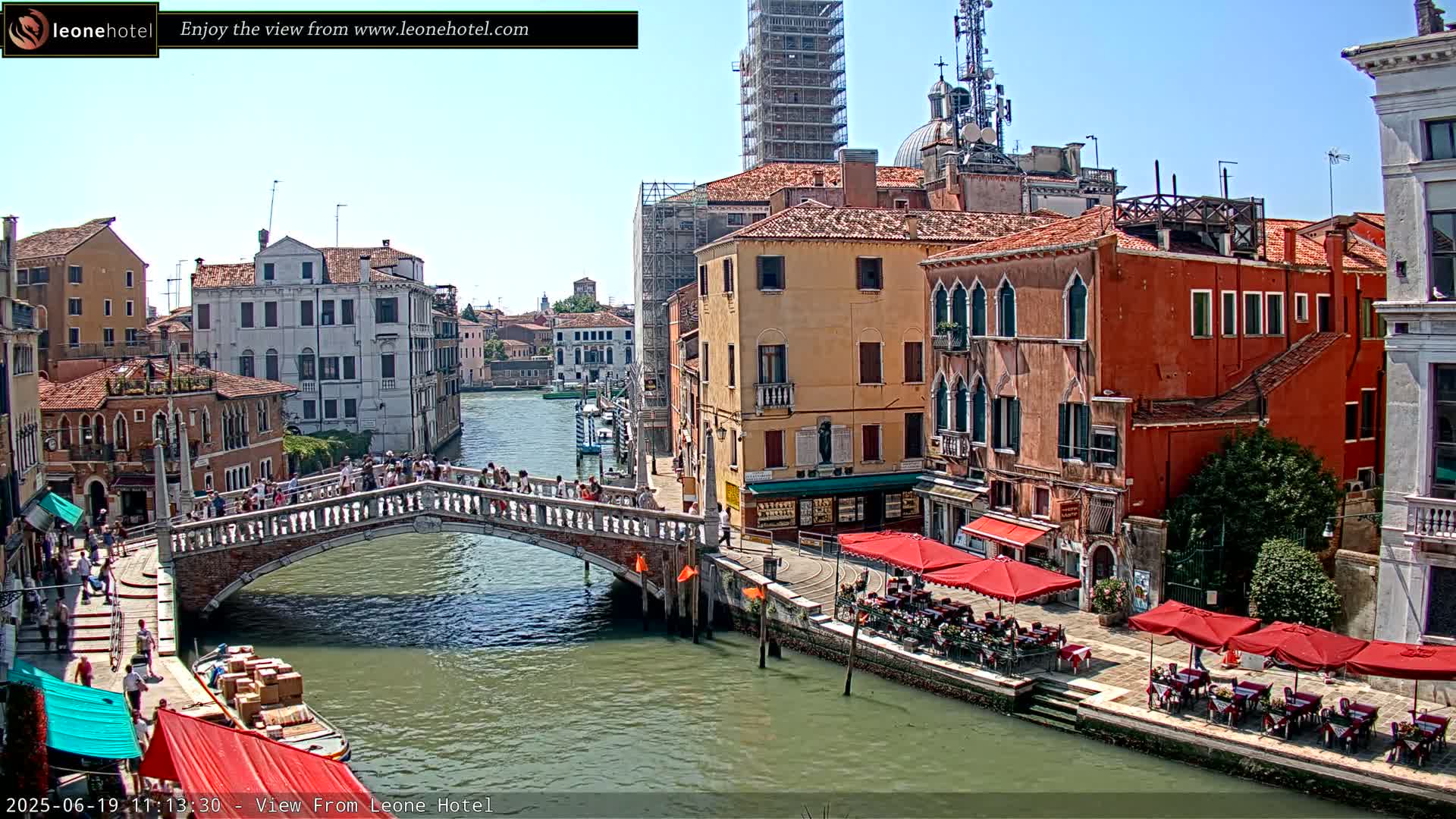 A sunny day in Venice shows a canal with a stone bridge, colorful buildings lining the waterways, and outdoor seating at cafes.