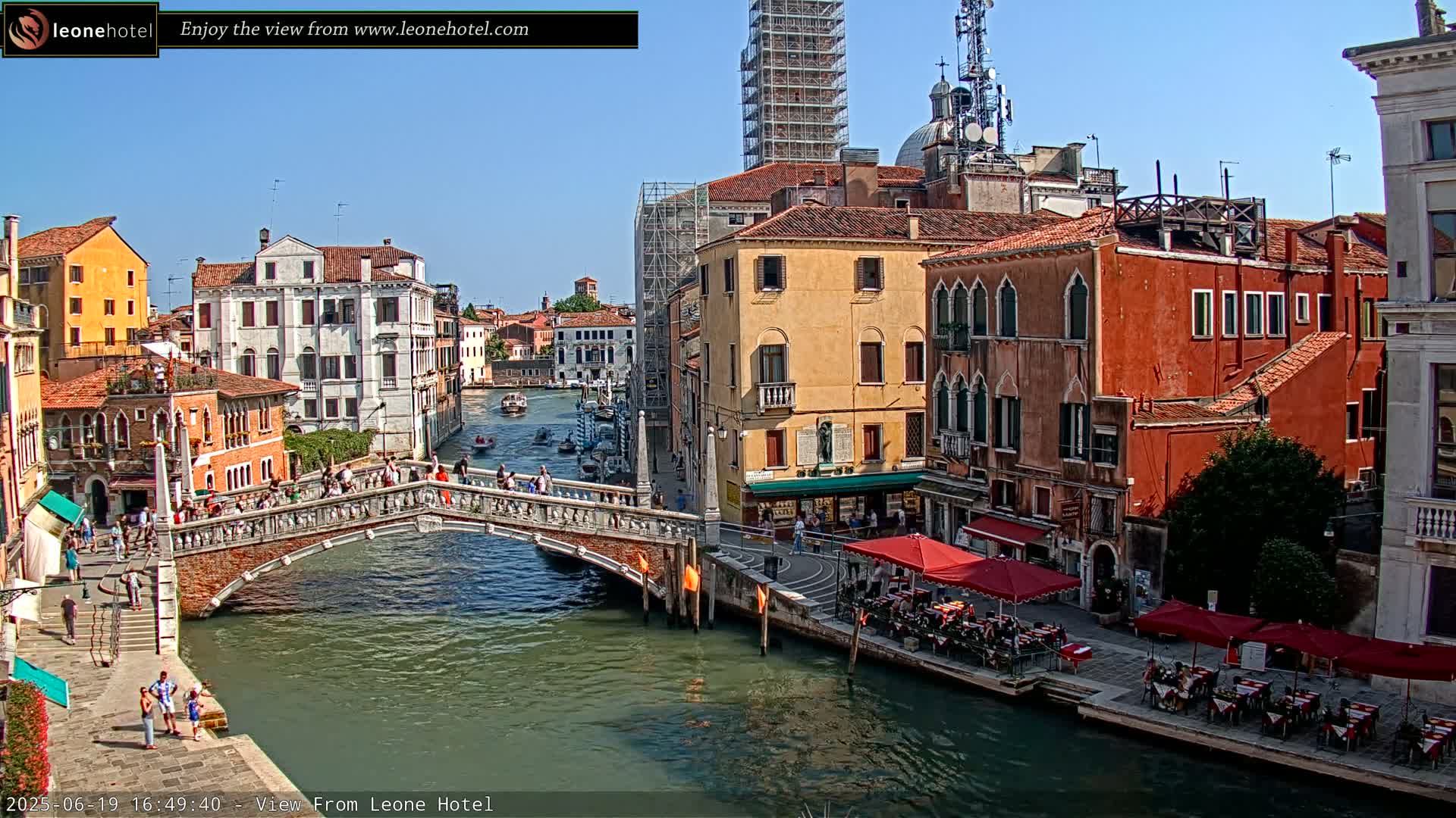 A sunny day in Venice shows a canal with a brick bridge, several colorful buildings lining the waterway, and outdoor seating at cafes along the canal.