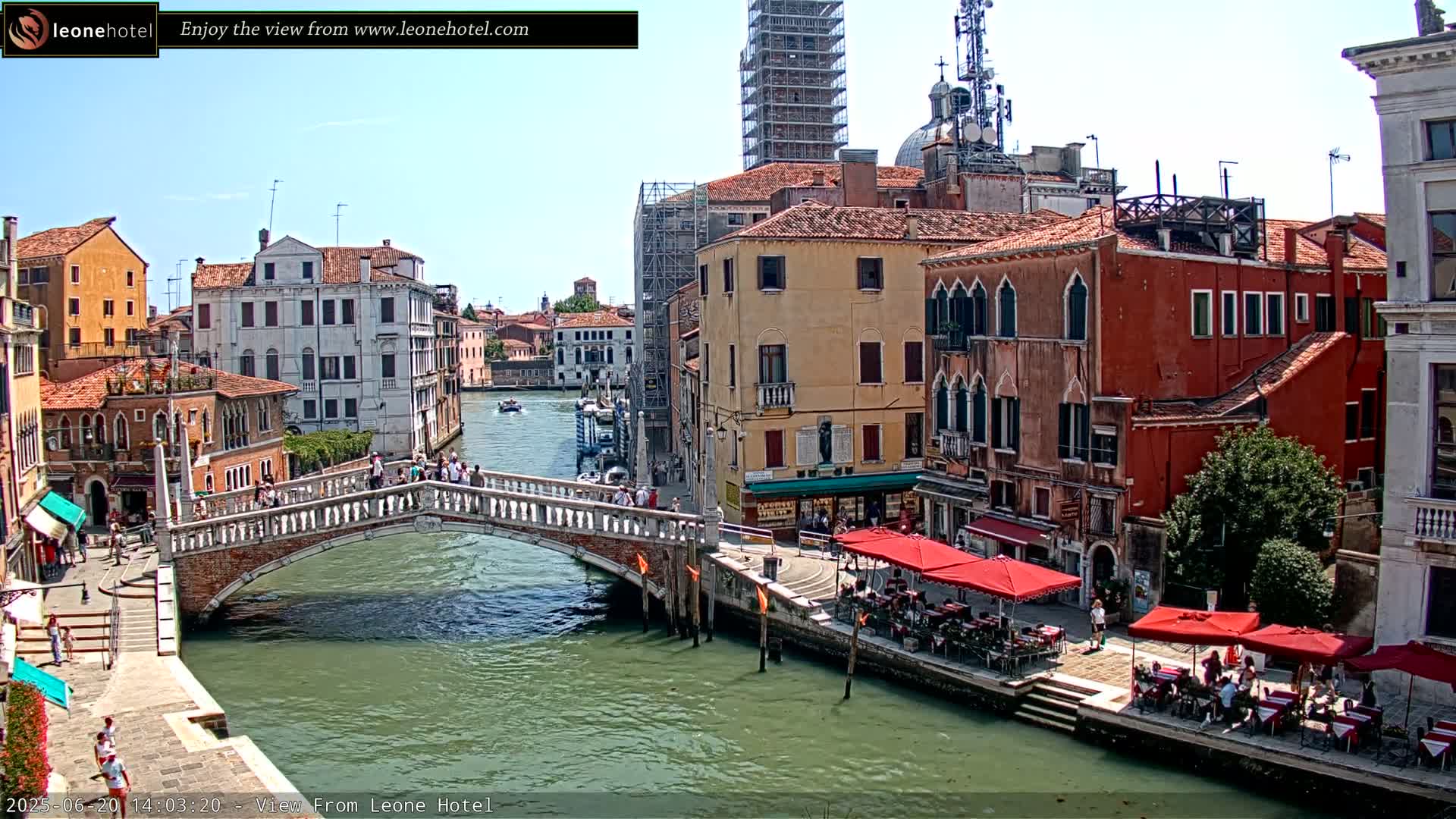 A sunny day in Venice shows a canal with a stone bridge, colorful buildings lining the waterway, and outdoor seating at cafes.