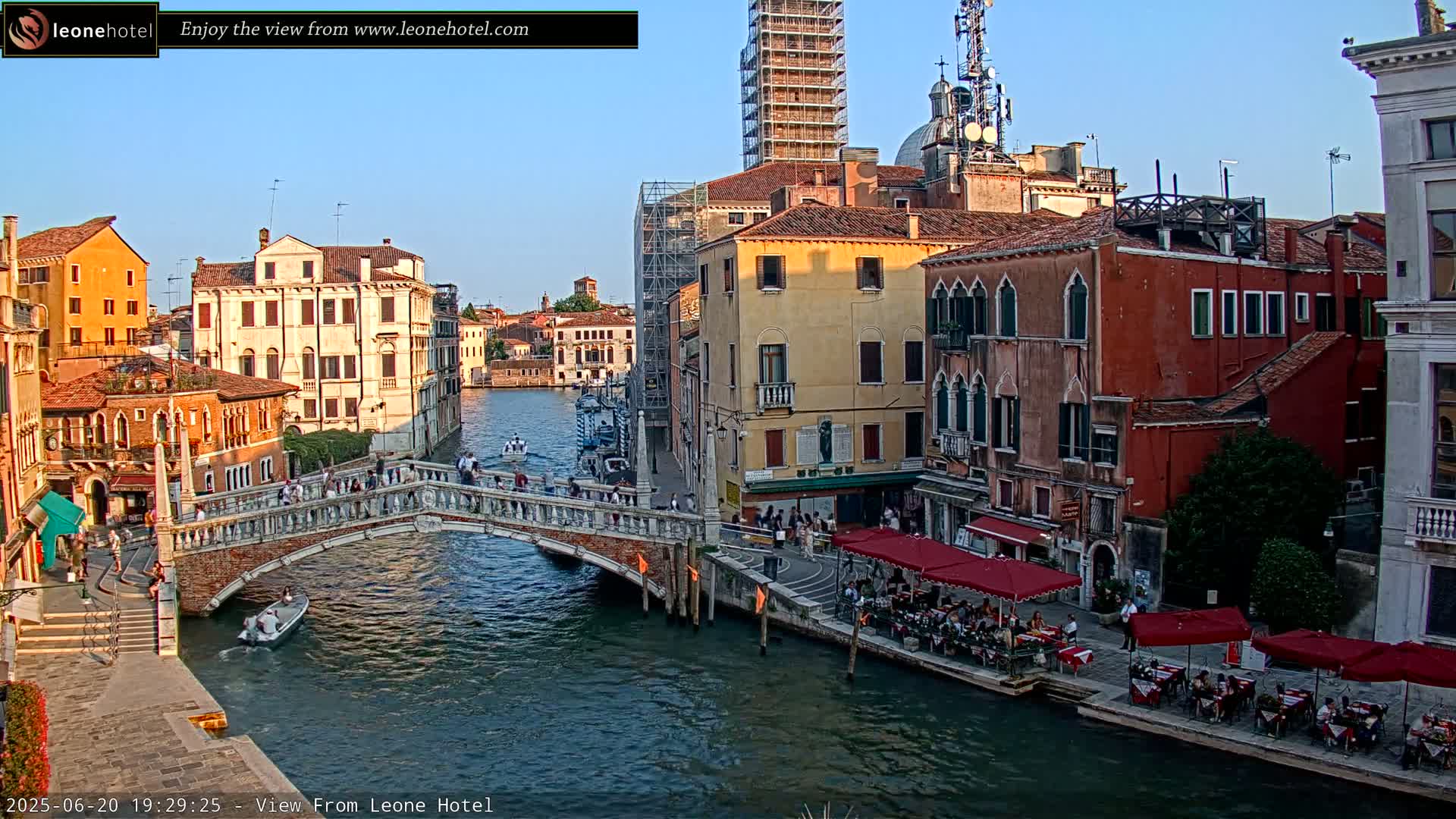 A stone bridge crosses a canal in Venice, Italy, under a clear evening sky, with colorful buildings lining the waterway and outdoor seating at restaurants along the canal.