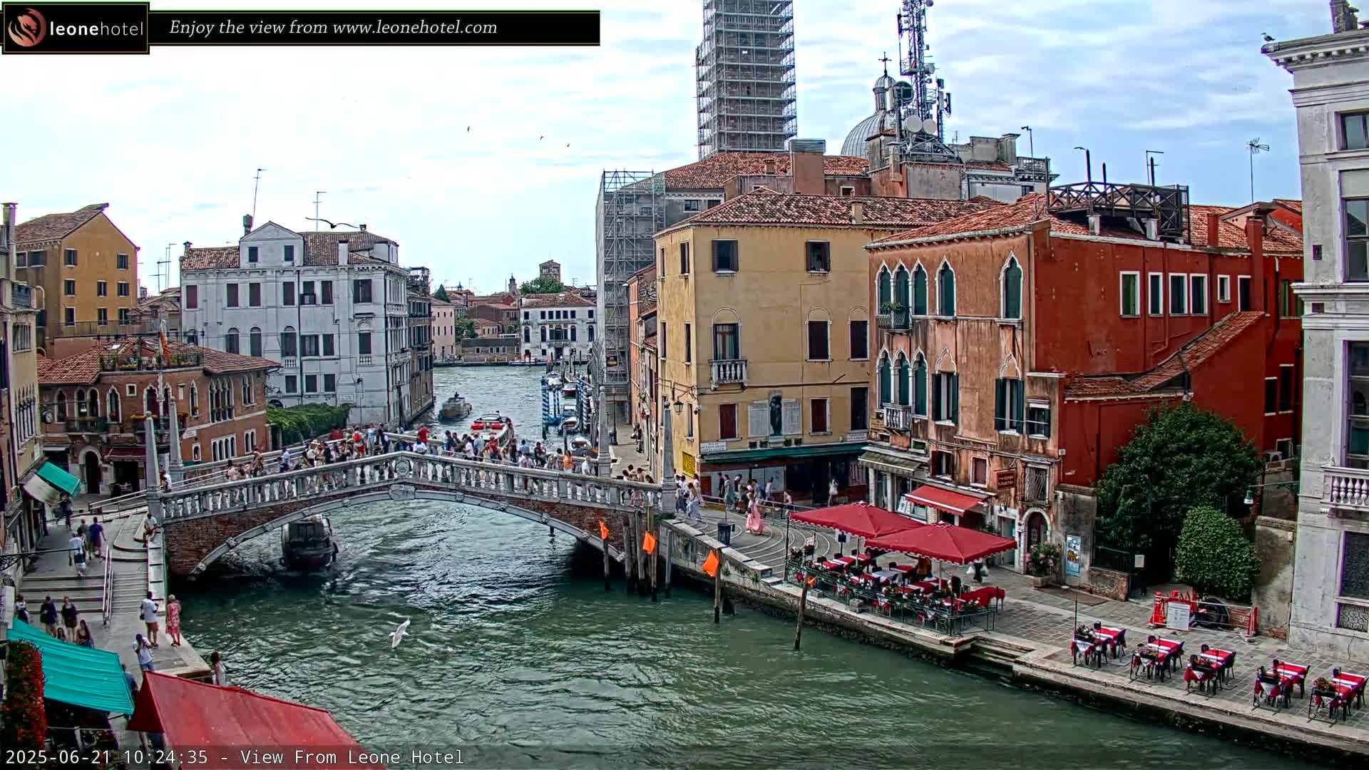 A bridge over a canal in Venice, Italy, is crowded with people, with colorful buildings and outdoor cafes lining the waterway under a partly cloudy sky.