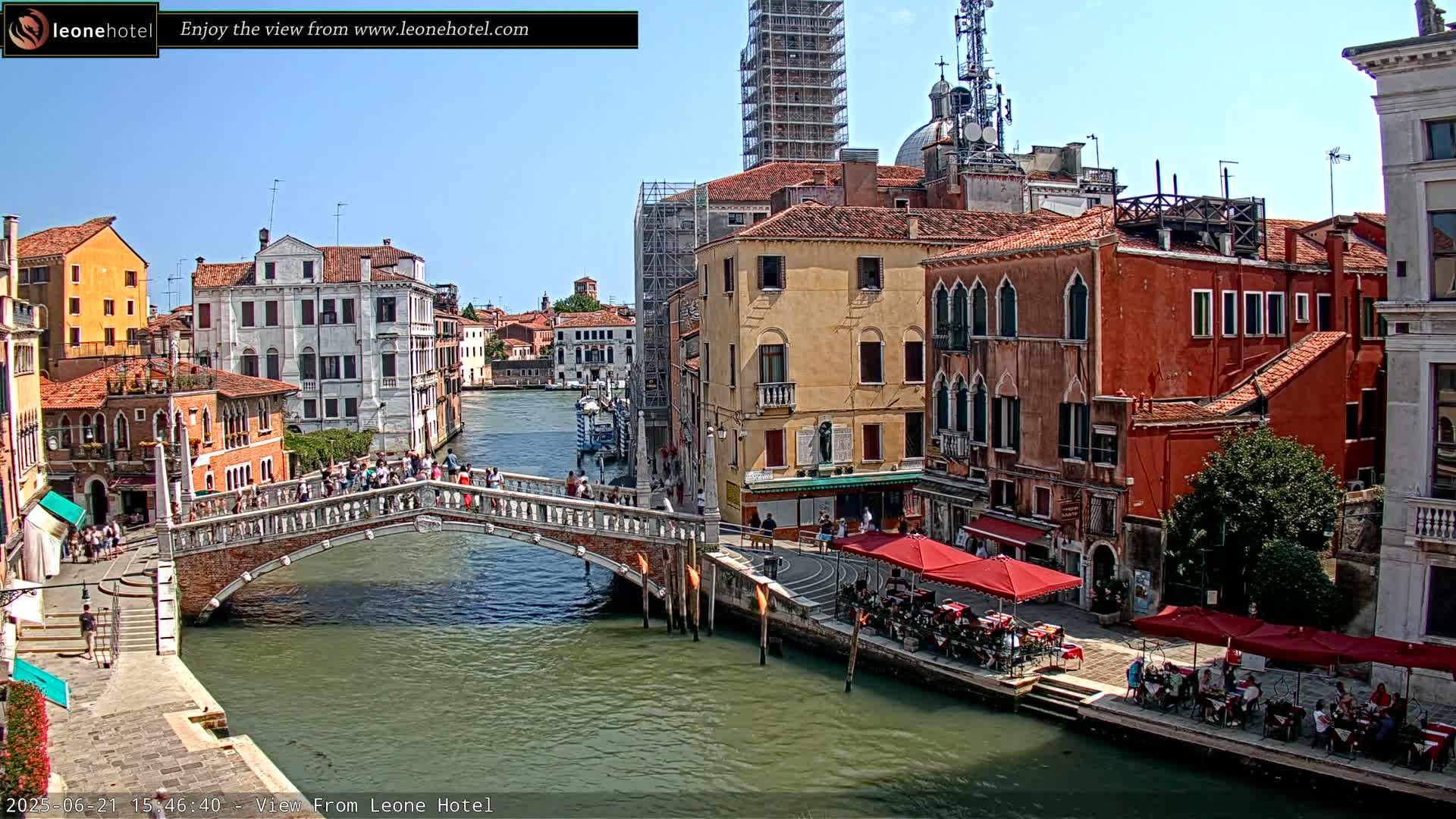 A bridge over a canal in Venice, Italy, on a sunny day, is surrounded by colorful buildings and people.