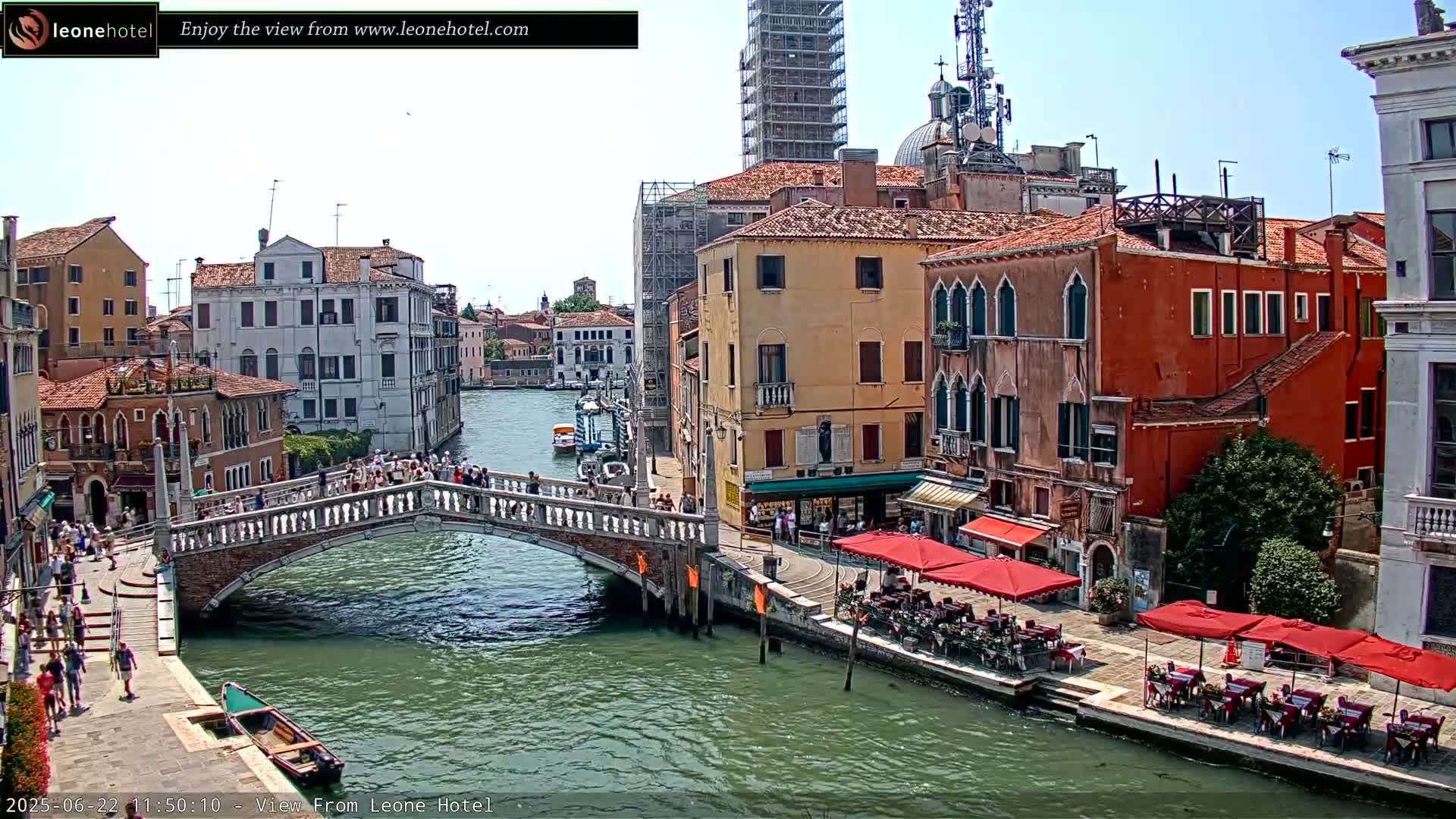 A sunny day in Venice shows a canal with a stone bridge, pedestrians, buildings, and outdoor seating at a cafe.