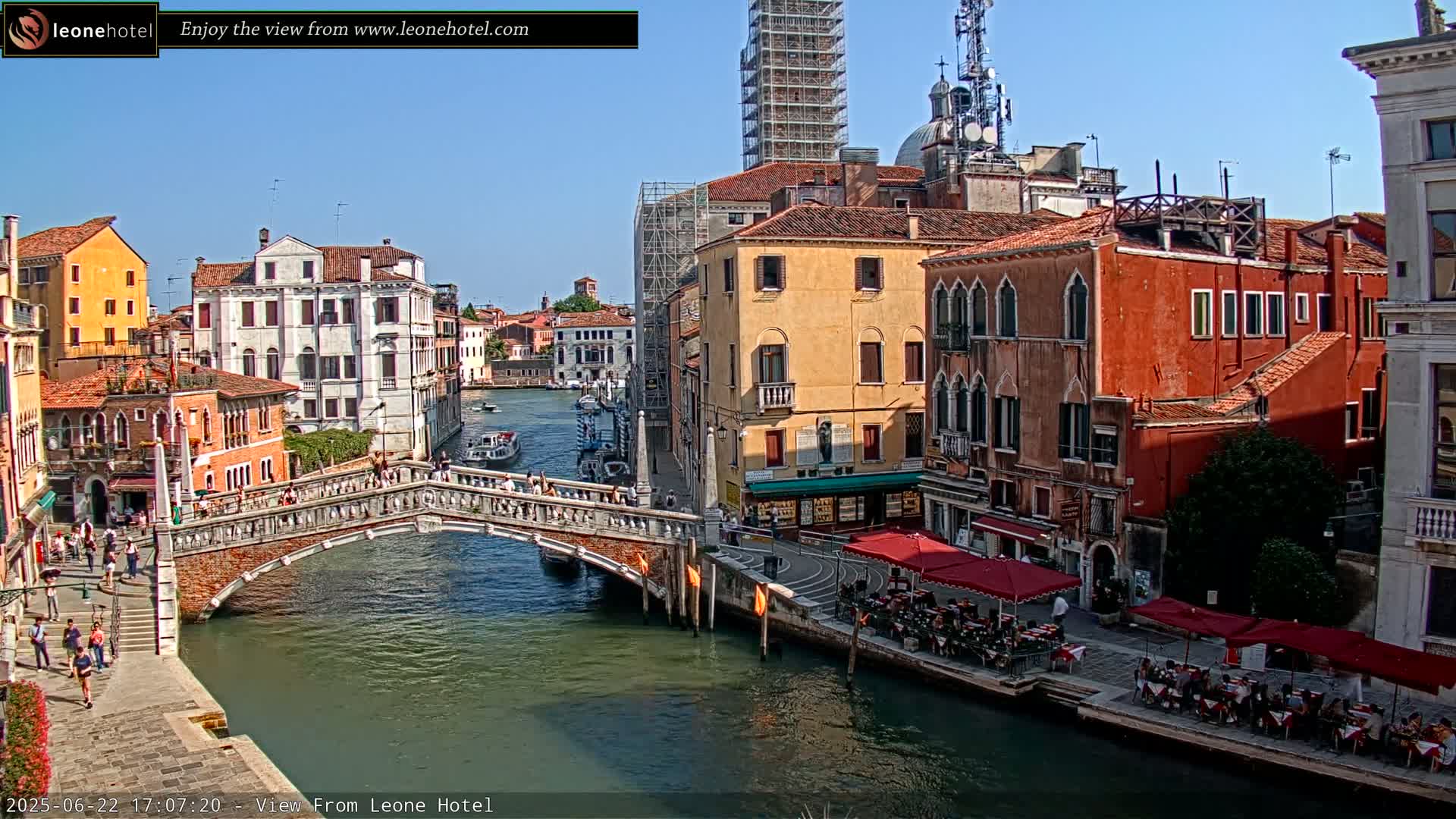 A sunny day in Venice, showing a canal with a stone bridge, colorful buildings lining the waterway, and outdoor seating at cafes.