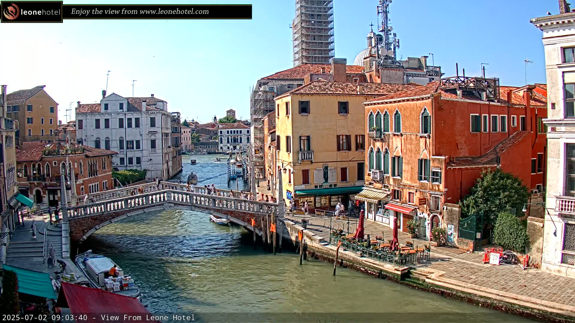 A sunny day in Venice shows a canal with boats, a stone bridge with people walking across it, and colorful buildings lining the waterway.