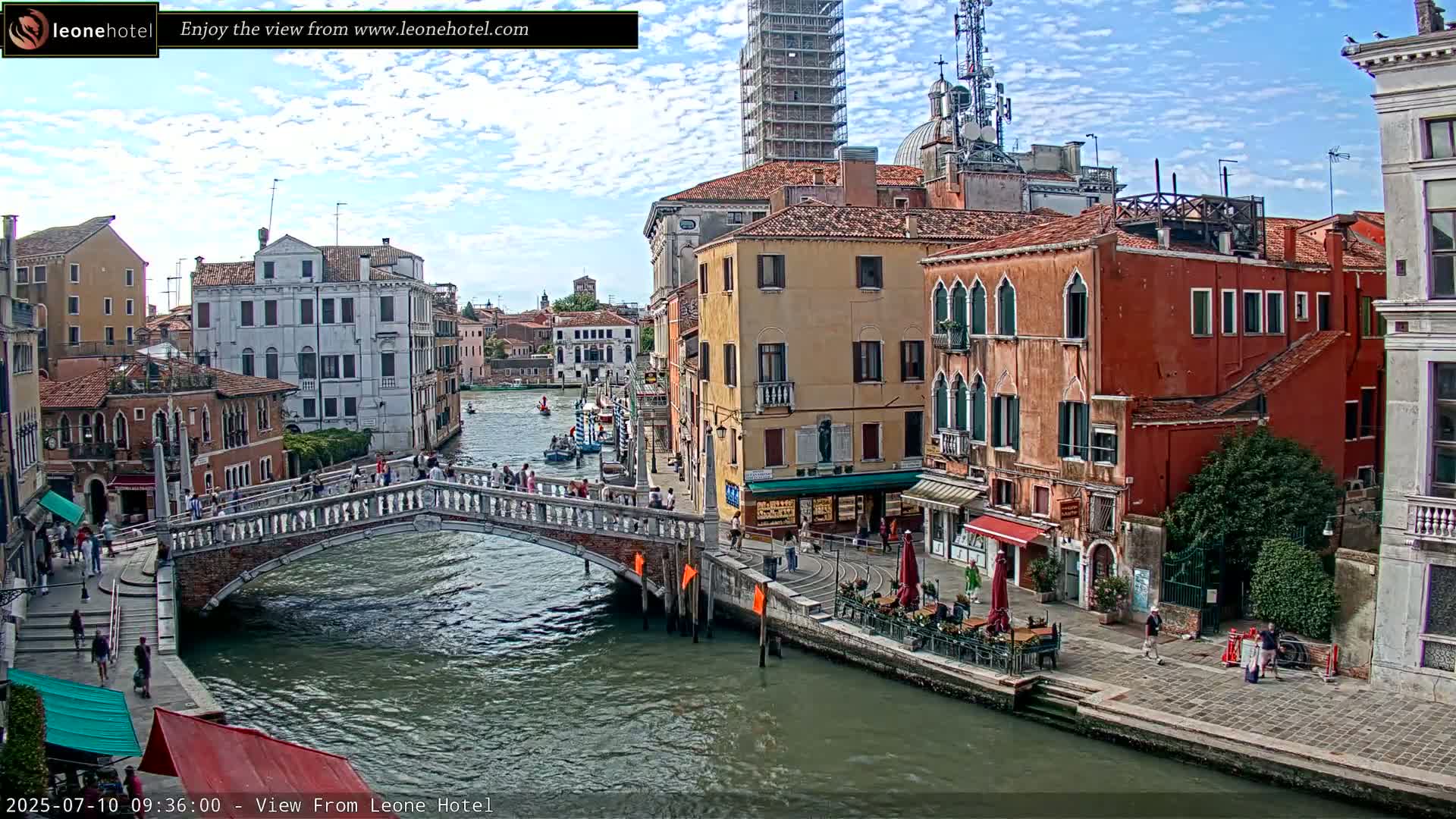 A bridge over a canal in Venice, Italy, under partly cloudy skies, with pedestrians strolling along the canal and buildings lining both sides.