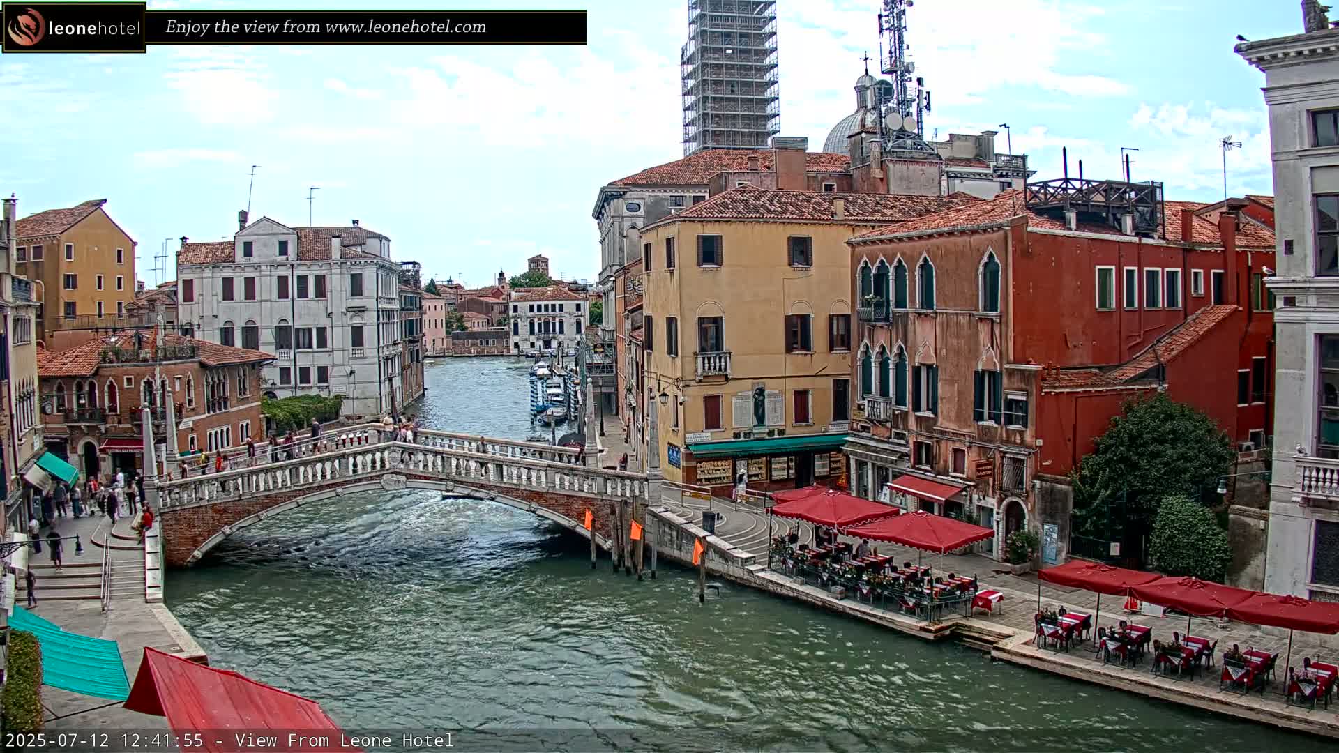 A bridge over a canal in Venice, Italy, under partly cloudy skies, with pedestrians strolling along the canal and buildings lining both sides.