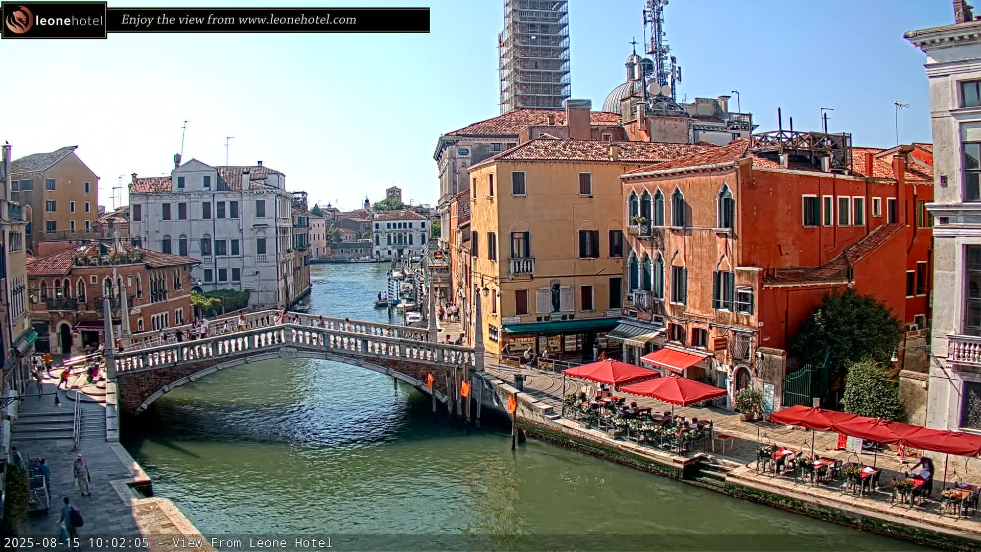 A sunny view of a canal in Venice, Italy, shows pedestrians crossing a stone bridge over the waterway lined with colorful buildings and outdoor cafes.
