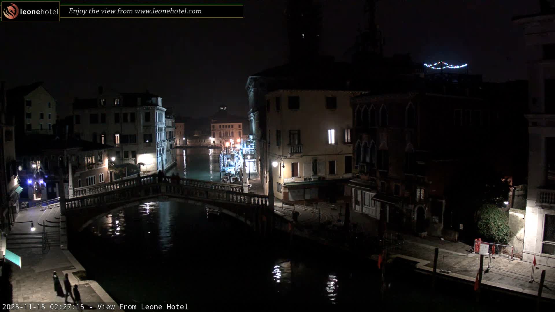 A nighttime view of a Venetian canal with illuminated bridges and buildings reflecting in the calm water under a clear sky.
