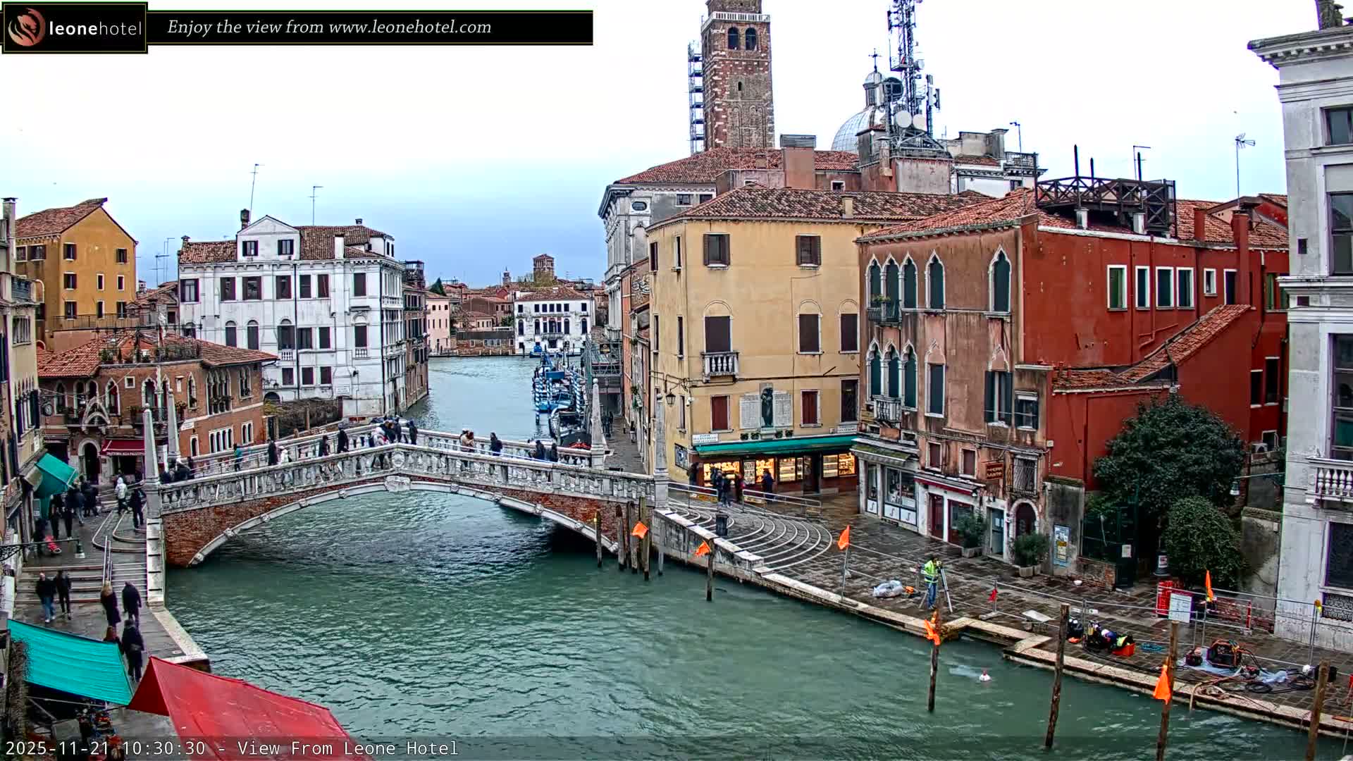 On a cloudy day in Venice, a picturesque canal winds through historic buildings, spanned by a stone bridge bustling with pedestrians, while workers attend to the waterfront.