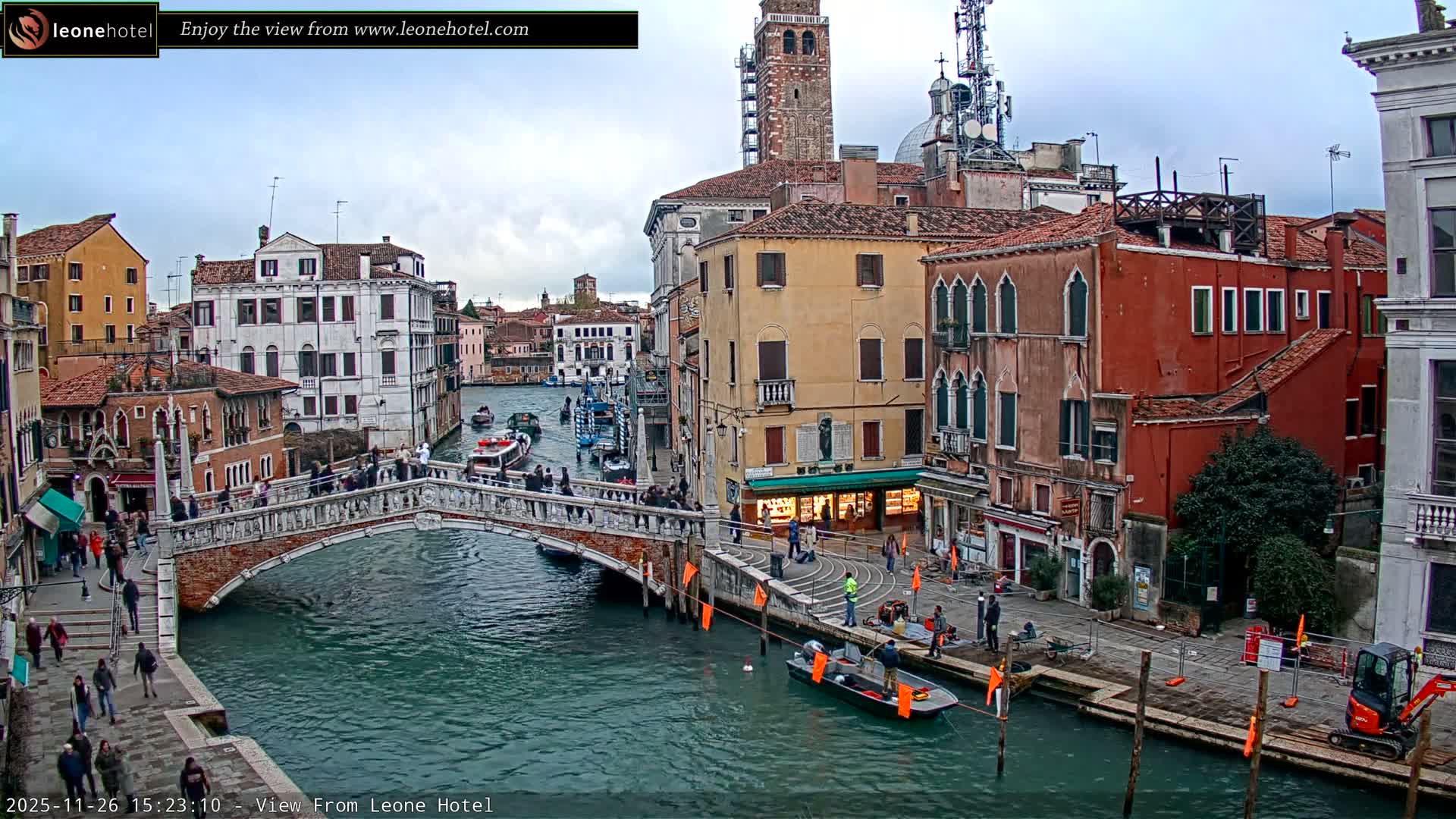 On a cloudy day in Venice, a picturesque canal winds through historic buildings, spanned by a stone bridge bustling with pedestrians, while workers attend to the waterfront.
