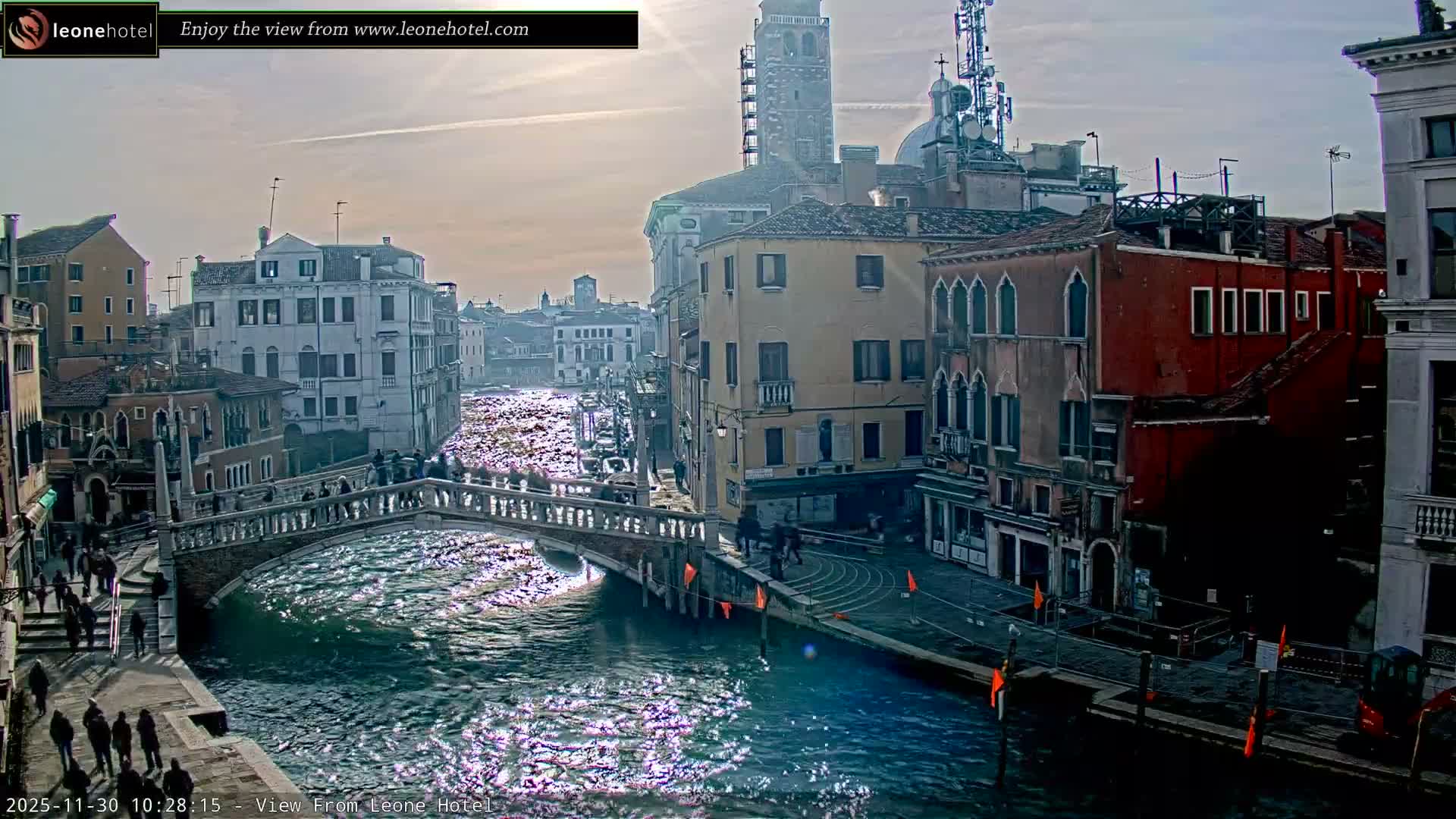 On a bright, sunny day, a bustling Venetian canal is visible, lined with historic buildings and crossed by a stone bridge, with people walking both on the bridge and along the water's edge.