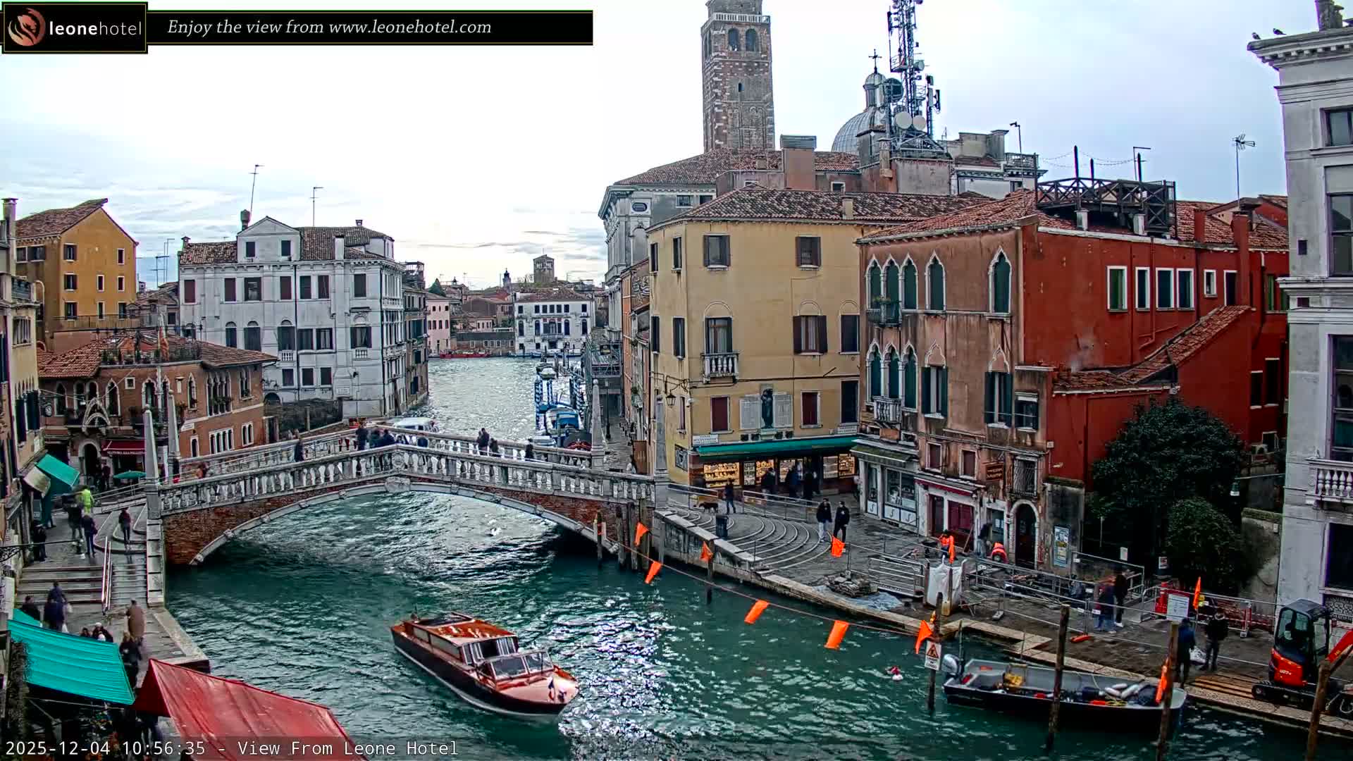 The image displays a bustling canal scene in Venice on an overcast day, featuring historic buildings lining the water, an ornate stone bridge with people crossing, a motorboat on the turquoise canal, and some construction activity visible on the right bank.