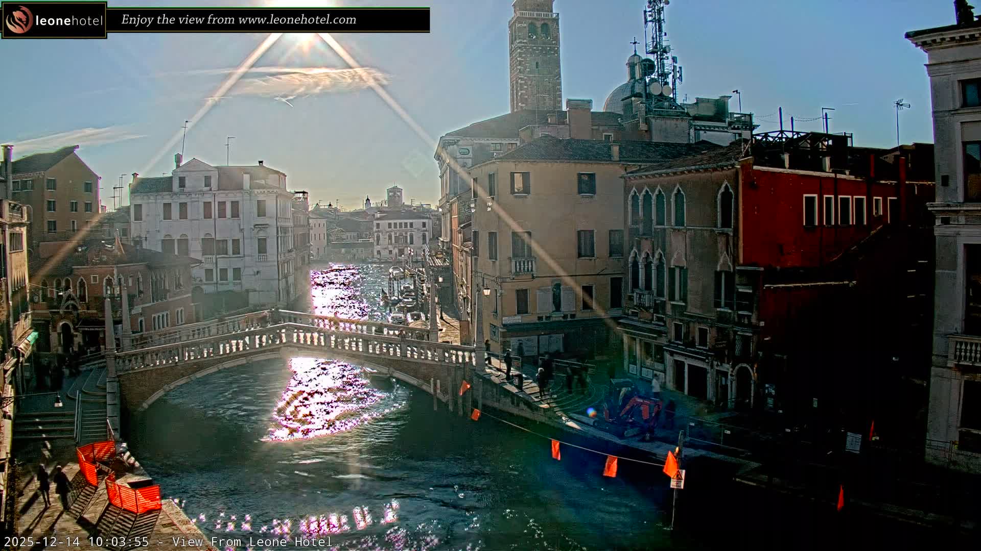 The image displays a bustling canal scene in Venice on an overcast day, featuring historic buildings lining the water, an ornate stone bridge with people crossing, a motorboat on the turquoise canal, and some construction activity visible on the right bank.