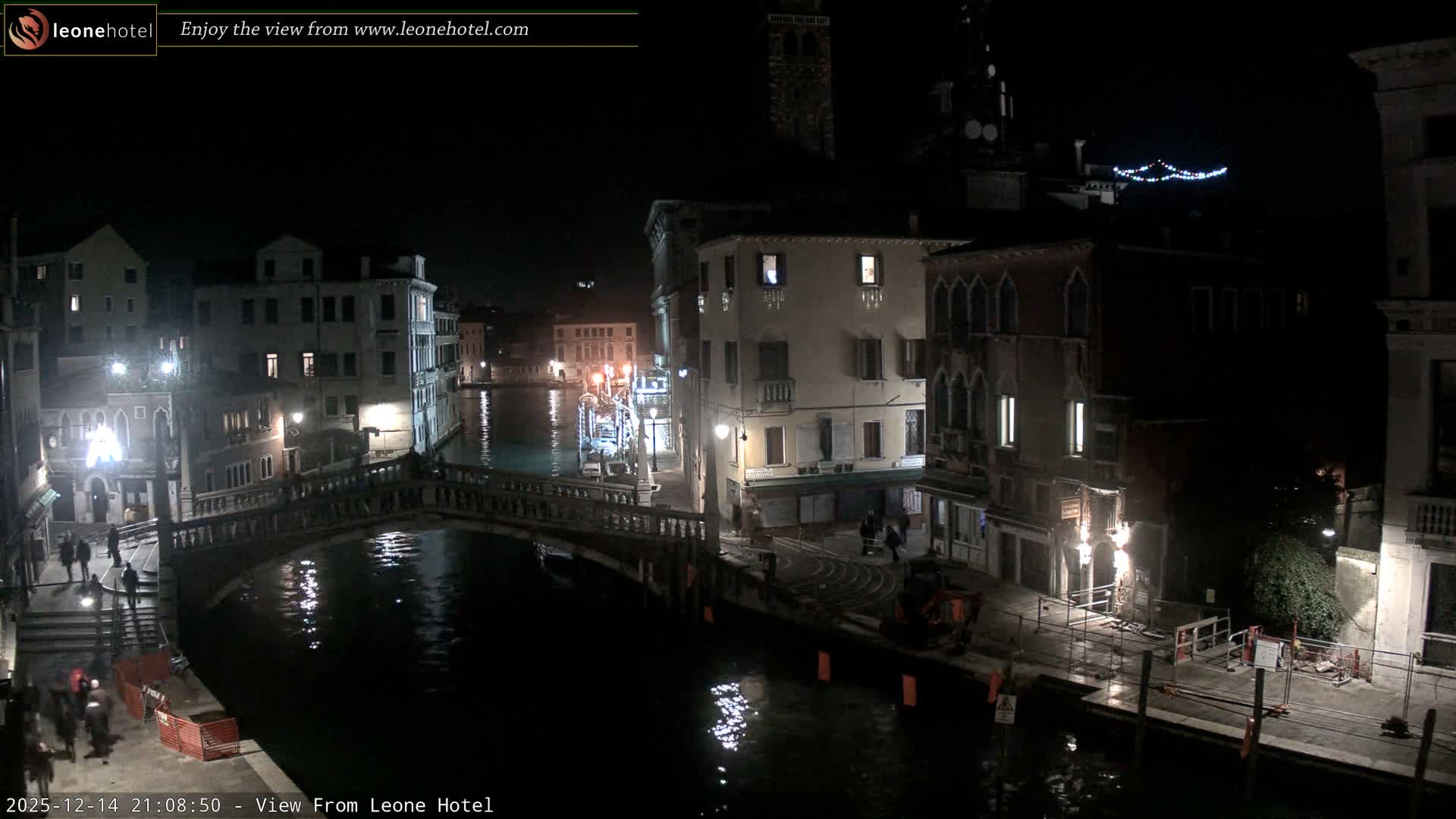 The image displays a bustling canal scene in Venice on an overcast day, featuring historic buildings lining the water, an ornate stone bridge with people crossing, a motorboat on the turquoise canal, and some construction activity visible on the right bank.
