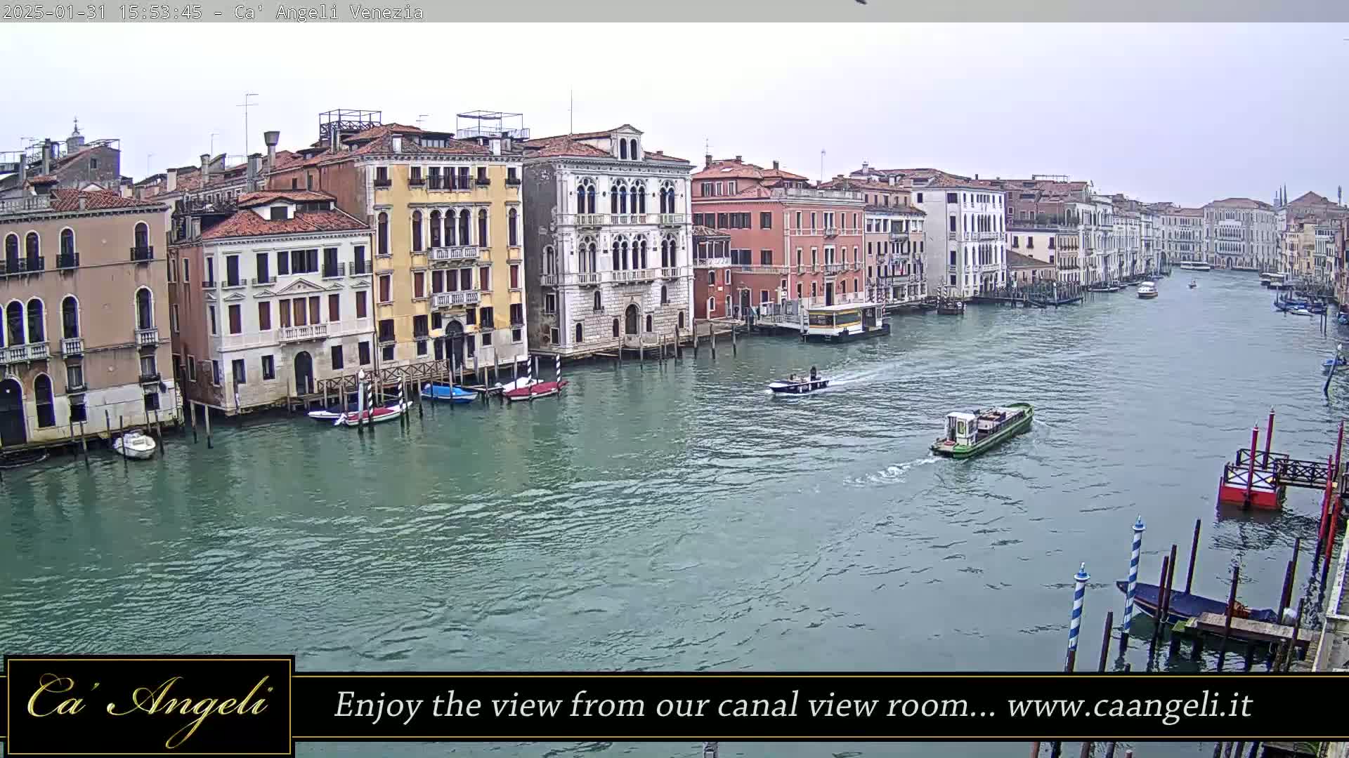 A wide shot of a canal in Venice, Italy, on an overcast day, shows numerous buildings lining the waterway and several boats traveling along it.