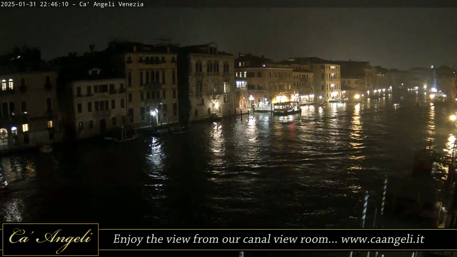 A nighttime view of a canal in Venice, showing illuminated buildings along its banks and boats on the water in rainy conditions.
