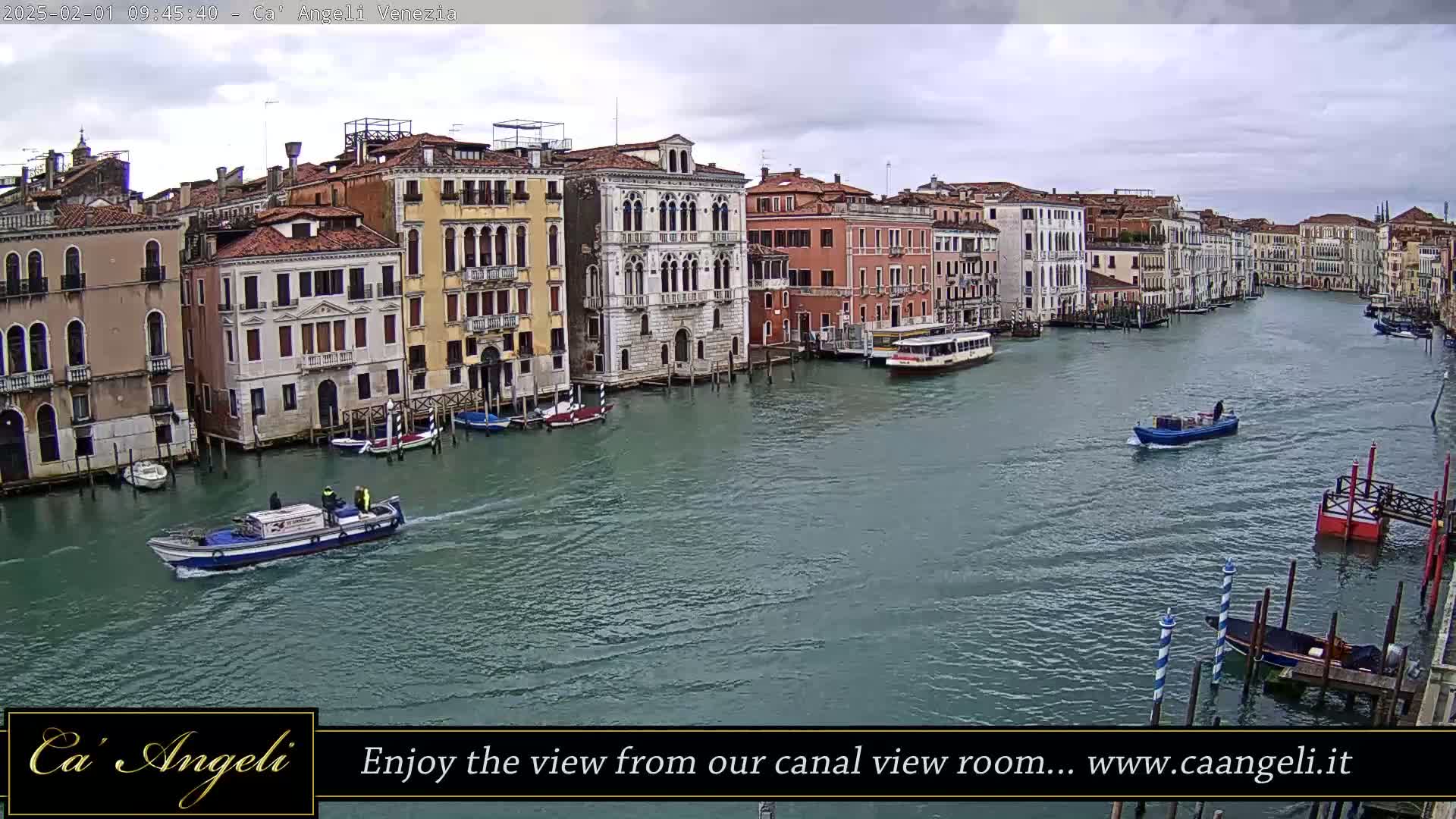 A wide shot of a canal in Venice, Italy, on an overcast day, shows several boats and numerous buildings lining the waterway.