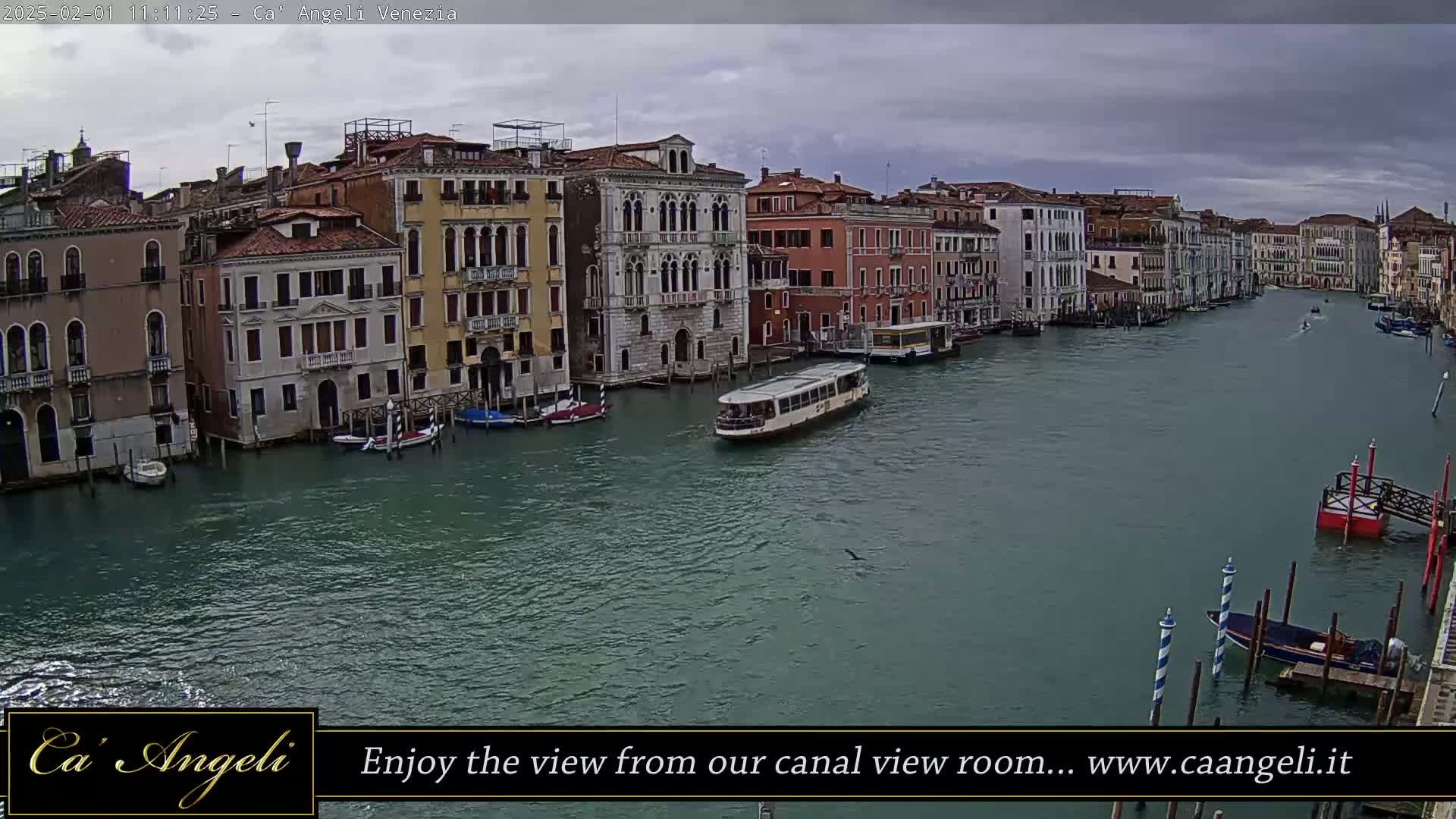 A waterway in Venice, Italy, shows several buildings along its banks under an overcast sky, with boats moored along the sides and one moving down the middle.