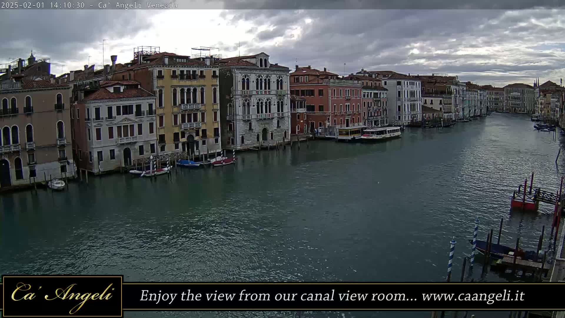 A wide shot of a canal in Venice, Italy, under a cloudy sky, shows several buildings lining the waterway with boats moored along the edge.