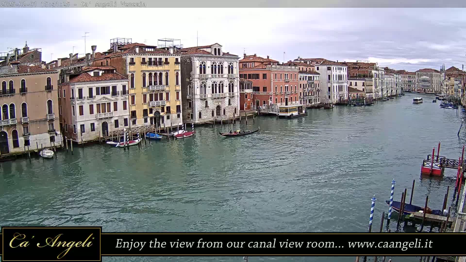 A wide shot of a canal in Venice, Italy, on an overcast day, shows several multi-story buildings lining the waterway, with boats and gondolas moored along the banks and navigating the channel.