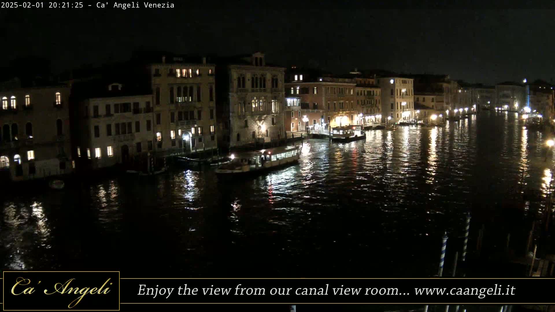 A nighttime view of a canal in Venice, Italy, shows several buildings lit up along the waterway, with boats moored along the banks and reflections shimmering on the dark water.