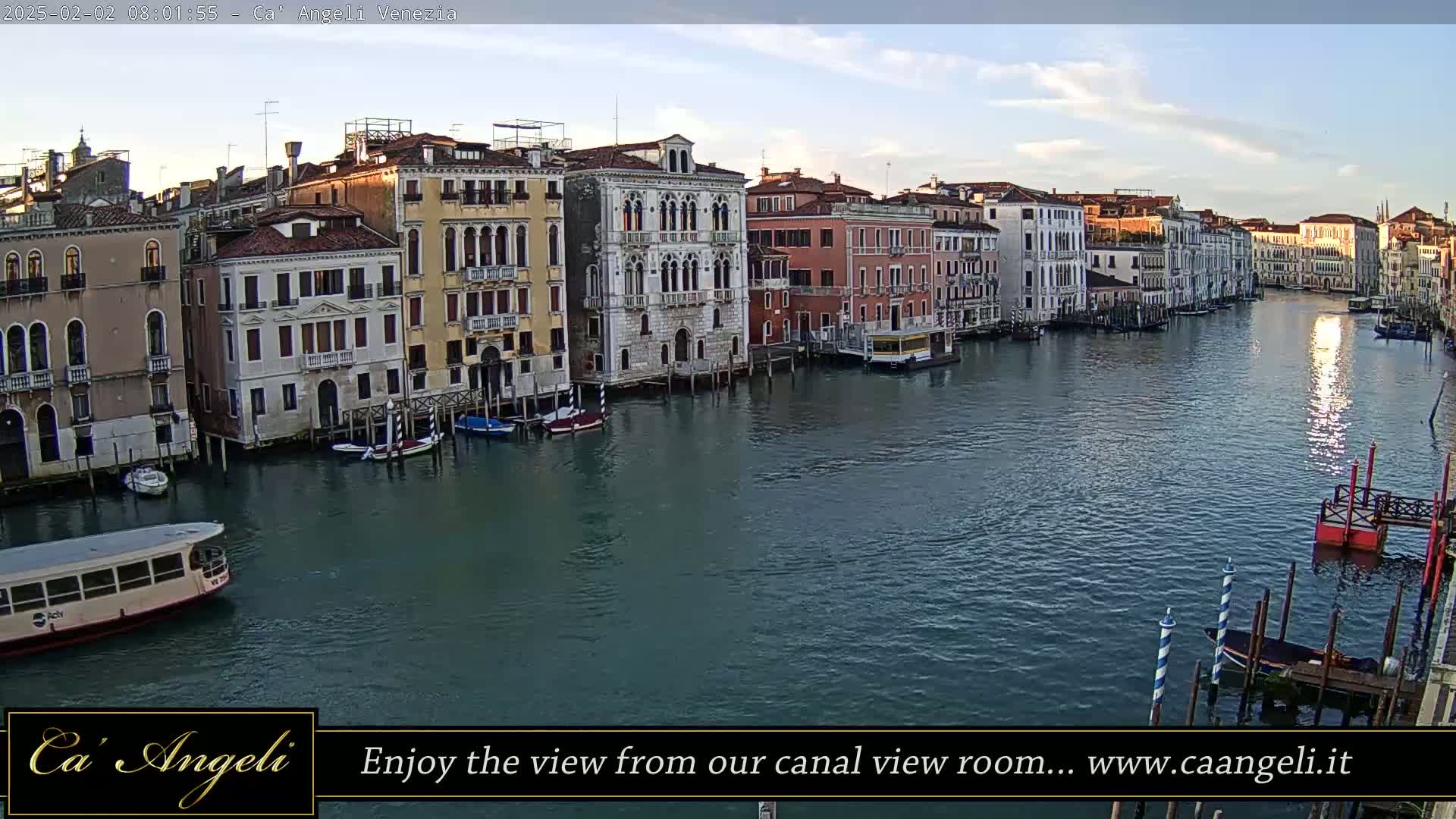 A wide shot of a Venetian canal at sunrise, showing several buildings along its banks and boats moored in the water.