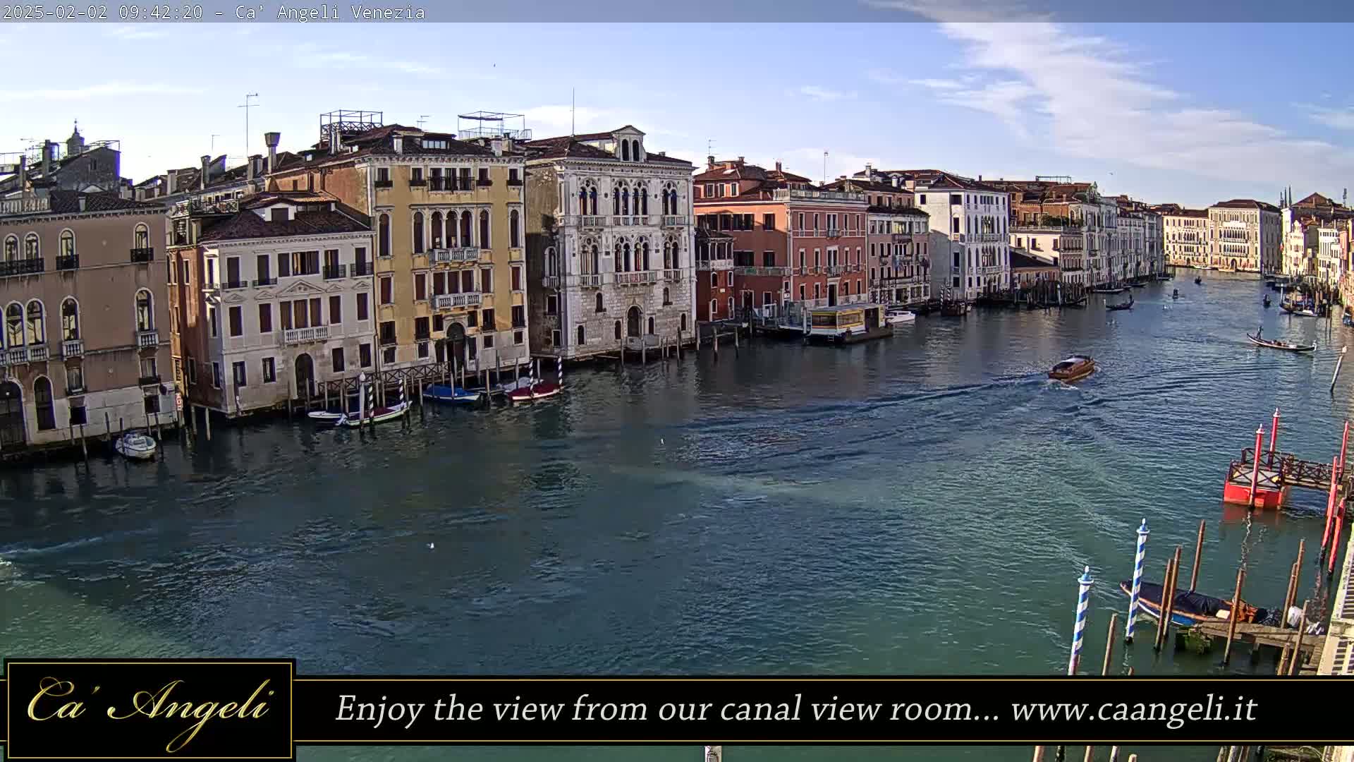 A sunny day in Venice shows a wide canal lined with historic buildings, several boats are visible on the water.