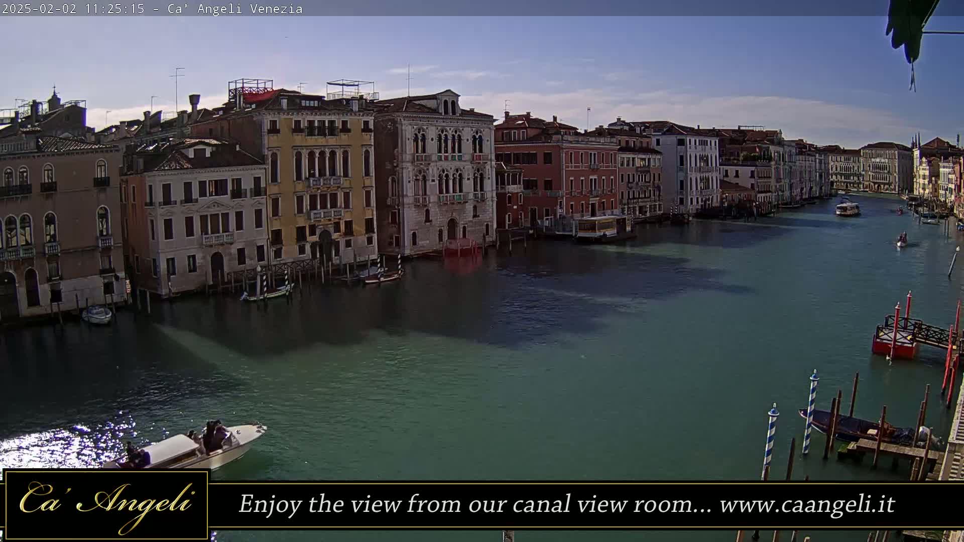 A wide shot of a waterway in Venice, Italy, shows several buildings along the shore on a sunny day, with boats on the water.
