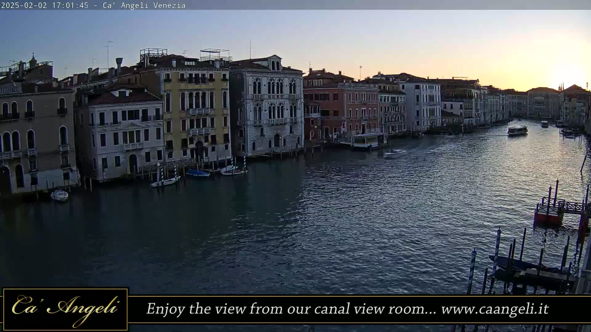 A twilight view of a canal in Venice, Italy, shows several buildings along its banks, with boats moored alongside and others traveling on the water.