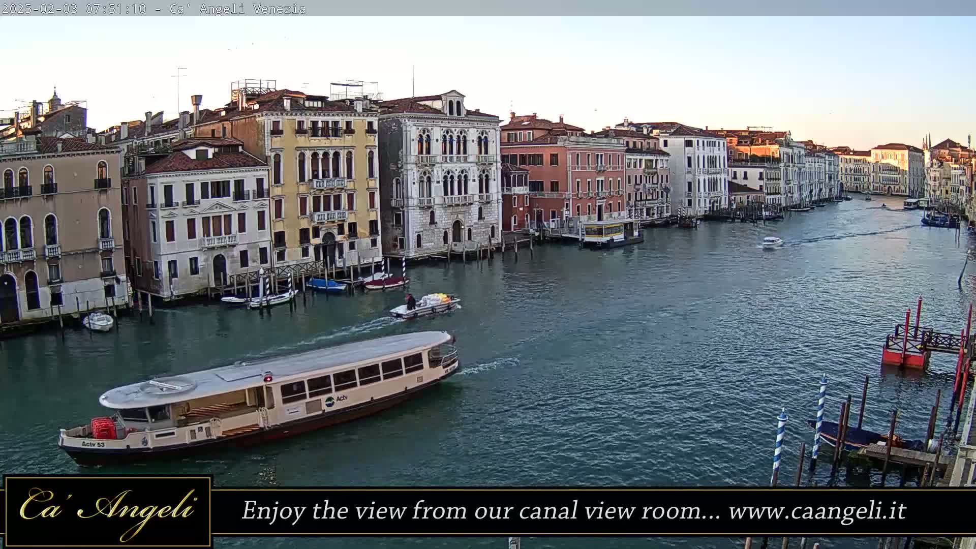 A wide canal in Venice, Italy, is shown on a calm, bright morning with several boats, including a passenger ferry, and many buildings lining the waterway.