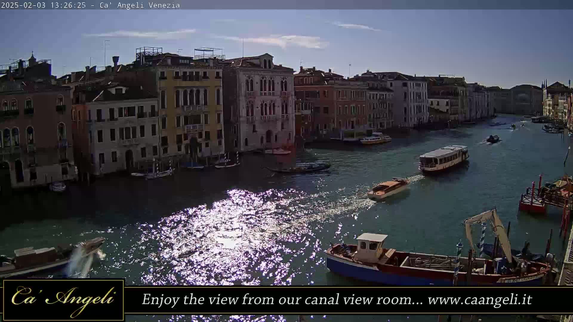 A sunny day in Venice shows a wide canal with several boats and gondolas passing by colorful buildings lining the waterway.
