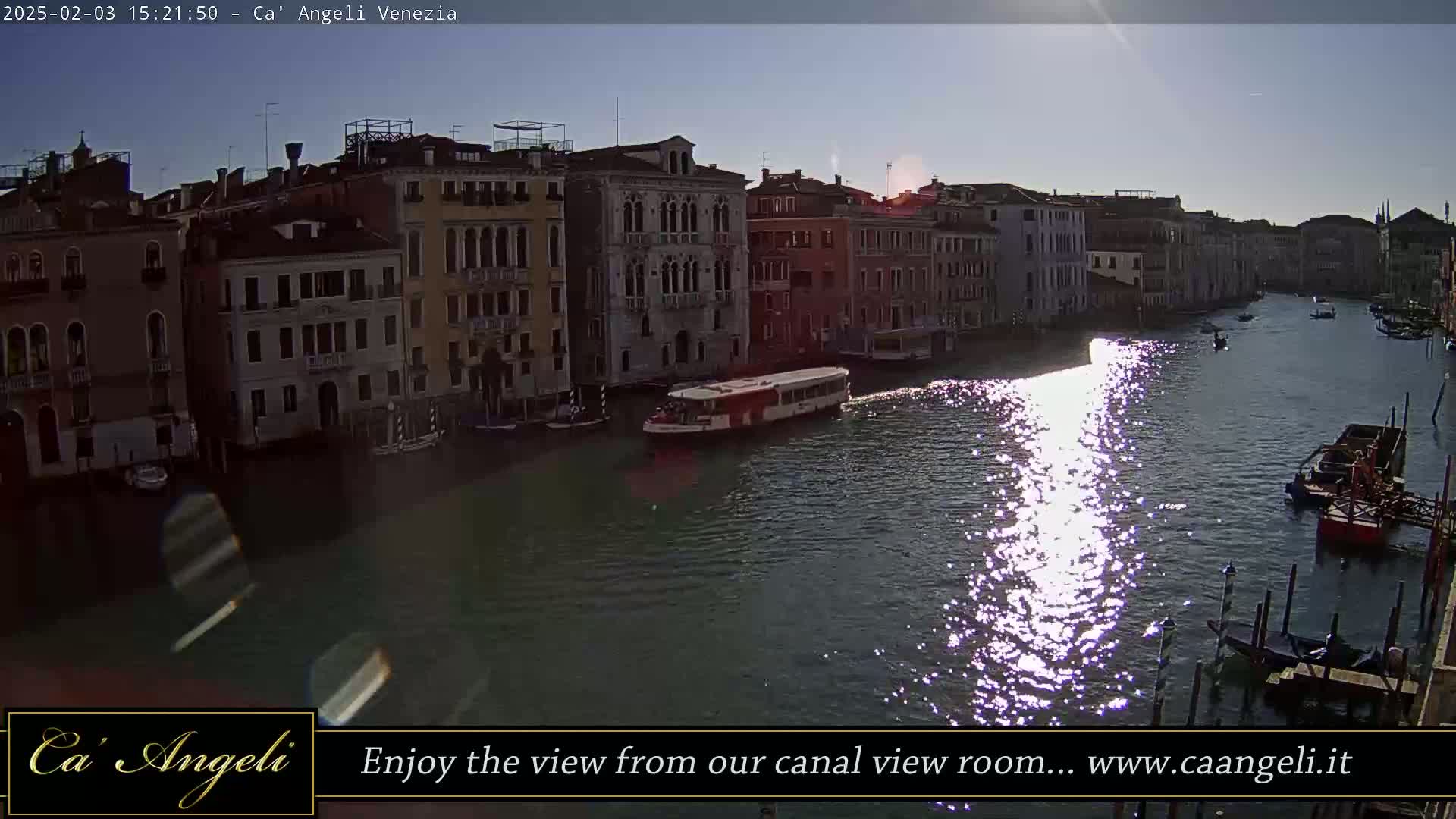A waterway reflecting sunlight, lined with tall buildings, with a few boats passing by under a mostly sunny sky.