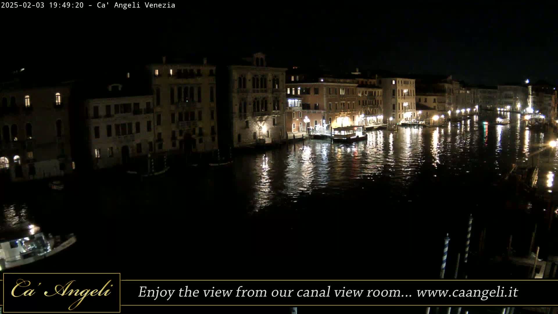 A nighttime view of a canal in Venice, showing illuminated buildings along the water's edge and boats on the calm water.