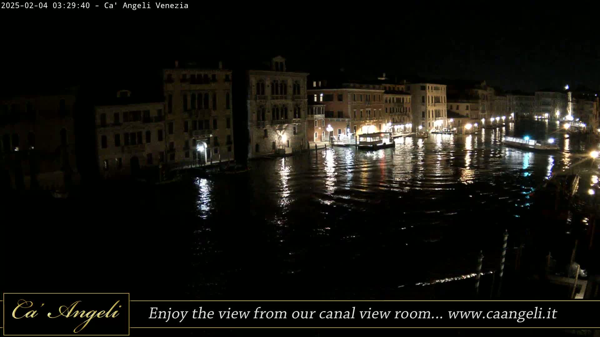 A nighttime view of a canal in Venice, Italy, shows buildings lining the waterway, with boats on the water and reflections in the dark water.