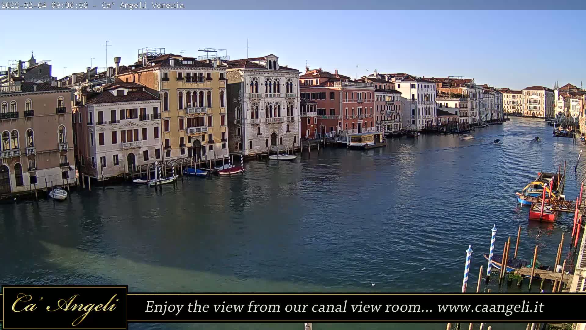 A wide canal in Venice, under a clear blue sky, is lined with historic buildings and several boats are moored along the waterway.
