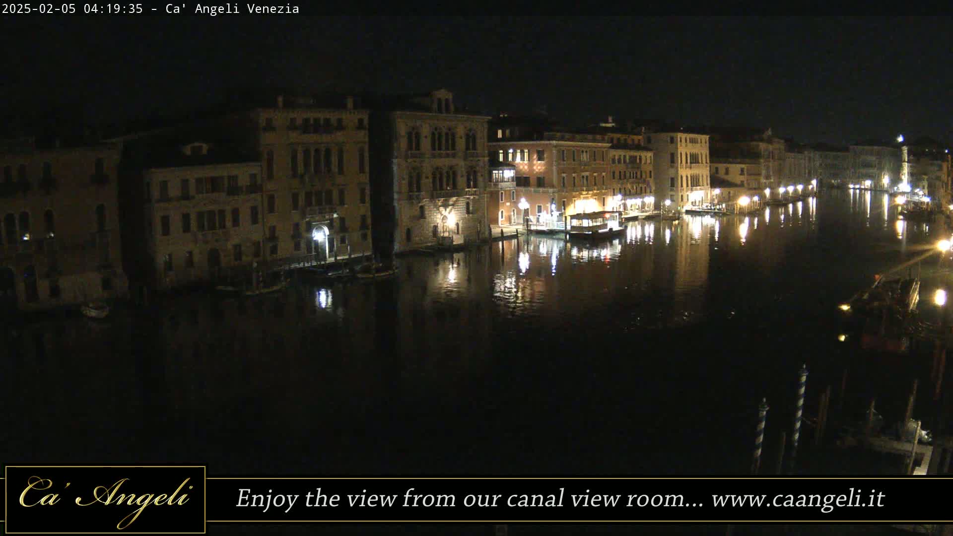 A nighttime view of a canal in Venice, lined with illuminated buildings and boats, reflecting in the dark water.